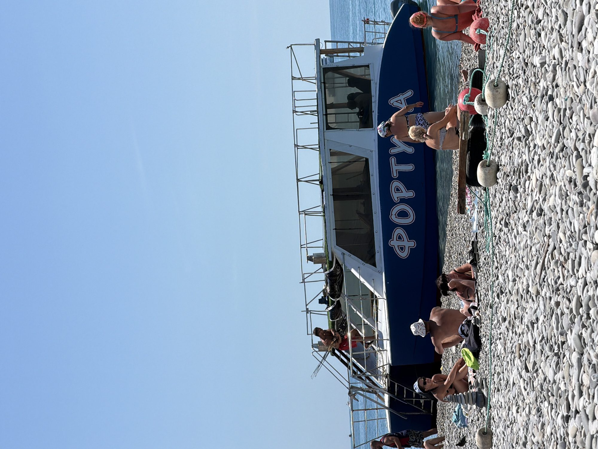On a sunny day, a pebble beach in Mamedova Shchel', Russia, is occupied by several people relaxing and engaged in various activities. A large blue and white catamaran, named "ФОРТУНА" (FORTUNA), is anchored in the shallow, calm sea, with some individuals on its deck and stairs. On the beach, a group of people are sunbathing, sitting, and lying on towels and the pebbles. One person is actively tending to fishing rods on the boat's deck. The environment is characteristic of a seaside resort, with clear blue skies and the sea meeting the shore.