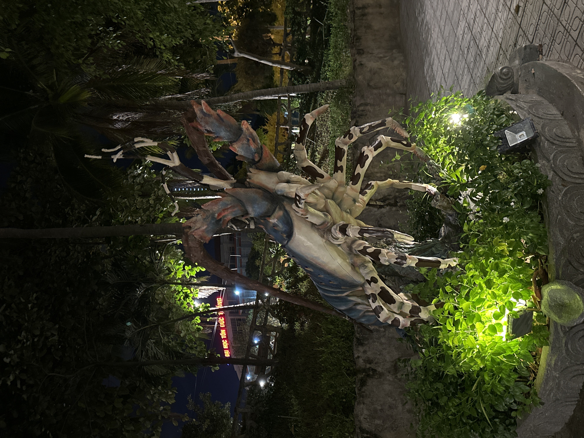 The image depicts a large, colorful sculpture of a lobster in an outdoor setting at night. The lobster sculpture is the central focus, with its elaborate claws and segmented body painted in shades of blue, white, and brown. It is situated amidst lush green foliage, which is illuminated by green-tinted lights, creating a dramatic effect. In the background, a building with red neon lettering that reads "ĐẠI HỌC TÔN ĐỨC THẮNG" (Ton Duc Thang University) is visible. This suggests the location is likely within or near the Ton Duc Thang University campus in Nha Trang, Vietnam, as the university has a campus in this city. Palm trees and other vegetation frame the scene, and a paved walkway with a distinctive pattern is visible to the right. The overall ambiance is somewhat surreal due to the nighttime lighting and the unusual subject matter of the sculpture.
