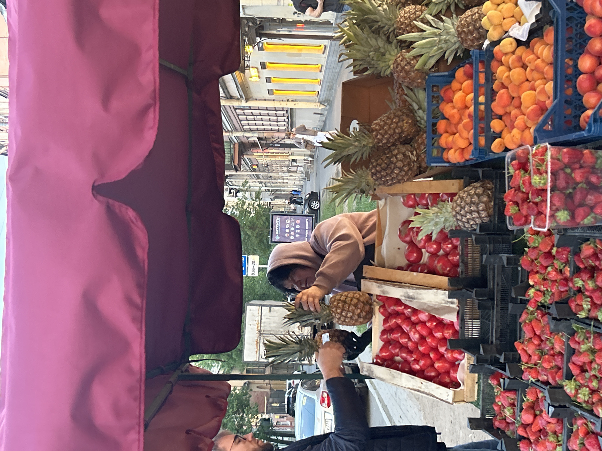 The image captures a vibrant street fruit market stall, viewed from a low angle, with a large red (magenta) canopy framing the foreground.
In the mid-ground, a person wearing a beige hoodie with black hair is seen arranging or selecting a pineapple from a display. Their hands are holding the fruit. In the immediate foreground, partially visible, another person with glasses and dark hair is looking towards the stall, with their arm holding a pineapple, suggesting they are either a customer or a vendor interacting with the fruit.
The market stall is abundant with fresh produce. On the right side of the image, numerous pineapples are stacked, some in wooden crates. Below them and across the stall, blue plastic crates are filled with bright orange apricots or peaches. Adjacent to these, clear plastic containers and black crates are overflowing with vivid red strawberries. Further left, crates of red apples or similar round red fruits are also visible.
In the background, a bustling street scene unfolds. Old buildings with traditional architecture line the road. A white car and a dark car are parked or driving further down the street. Several pedestrians are visible in the distance.
Notable details include a small sign with operating hours "8.00 - 20.00" (8 AM to 8 PM) and a digital screen displaying various icons and partially readable Cyrillic text (e.g., "ВМЕСТЕ" - "together," and what appears to be "ВЕТЧИНА" - "ham/bacon," possibly indicating other offerings or a generic template). The lighting suggests natural daylight, likely daytime on a clear day. The presence of Cyrillic text and the architectural style are consistent with the location context of Saint Petersburg, Russia. The overall scene depicts a typical urban market exchange.