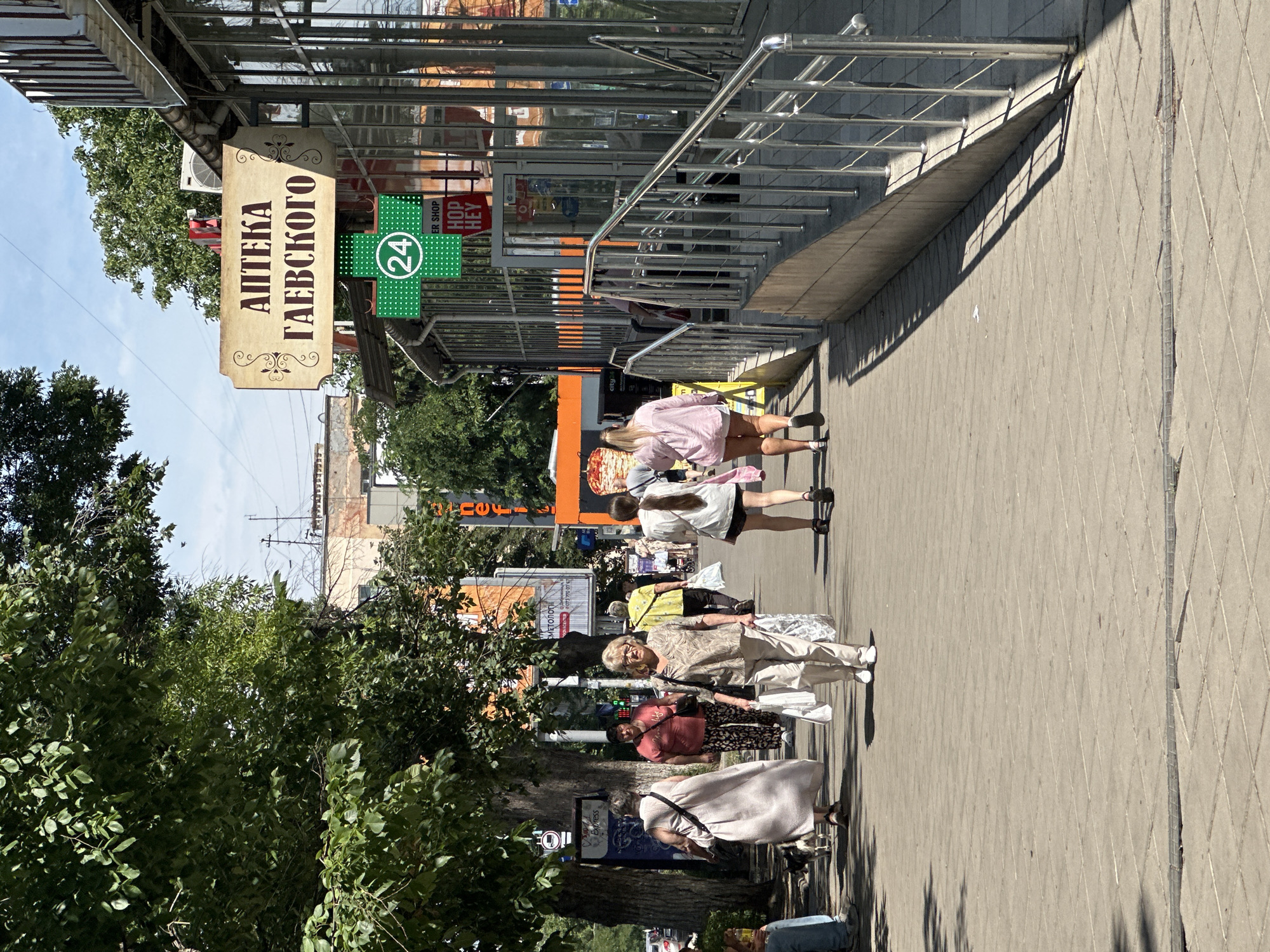 The image depicts a street scene in Odesa, Ukraine, on a sunny day. A paved sidewalk lines the right side of the image, leading past a building with large glass windows. A handrail guides a ramp descending from the sidewalk to a lower level. A sign above the building reads "АПТЕКА ГАЕВСКОГО" (GAEVSKY PHARMACY) in Cyrillic. Below it, a green sign with a white "24" and a cross indicates a 24-hour pharmacy. Several people are walking on the sidewalk. In the foreground, a woman in a light-colored outfit walks towards the camera, carrying shopping bags. To her left, another woman in a long, light-colored dress walks away from the camera, accompanied by a small dog. Further back, more pedestrians are visible, some carrying bags. Lush green trees overhang the sidewalk, casting shadows on the pavement. The overall impression is a typical urban street with commercial activity and pedestrian movement.