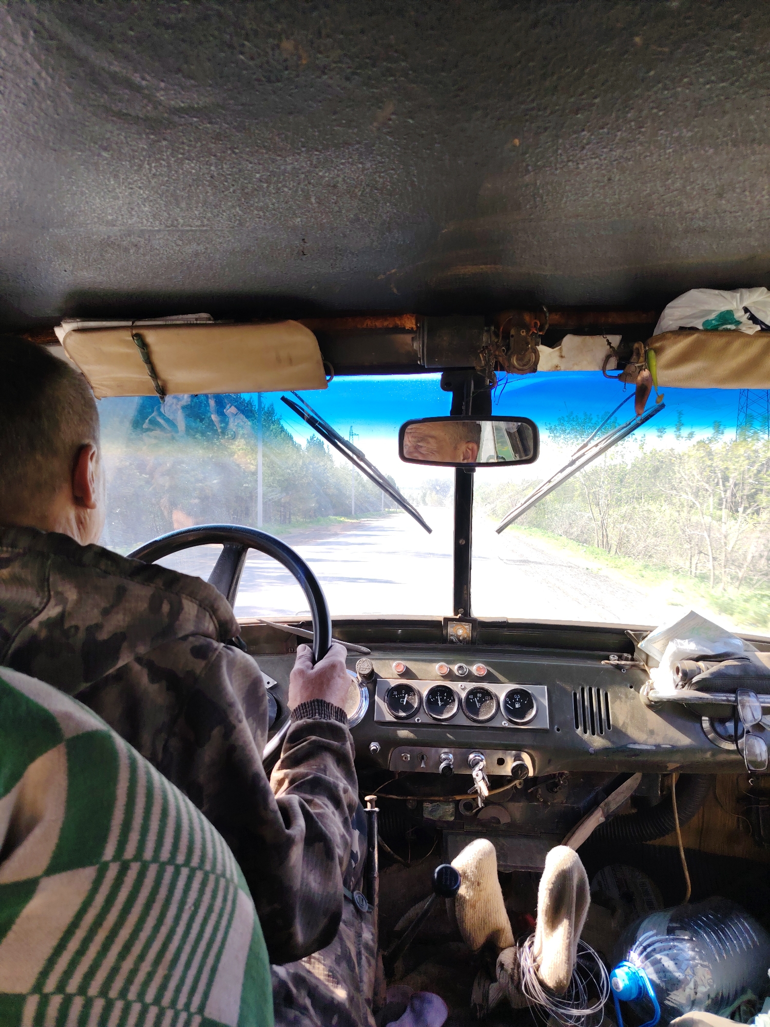 A man is driving an older vehicle, likely a UAZ, on a paved road. The scene is viewed from inside the vehicle, looking through the windshield. The driver is wearing a camouflage jacket and has short hair. His hand is on the steering wheel. The dashboard of the vehicle is visible, with gauges and controls. There are various items scattered around the interior, including a water bottle, socks, and possibly maps or documents. The road ahead is straight and lined with trees and power lines. The reflection in the rearview mirror shows the driver's face. The location may be in the Chaikovsky, Russia area, based on the likely type of vehicle and the surrounding environment.