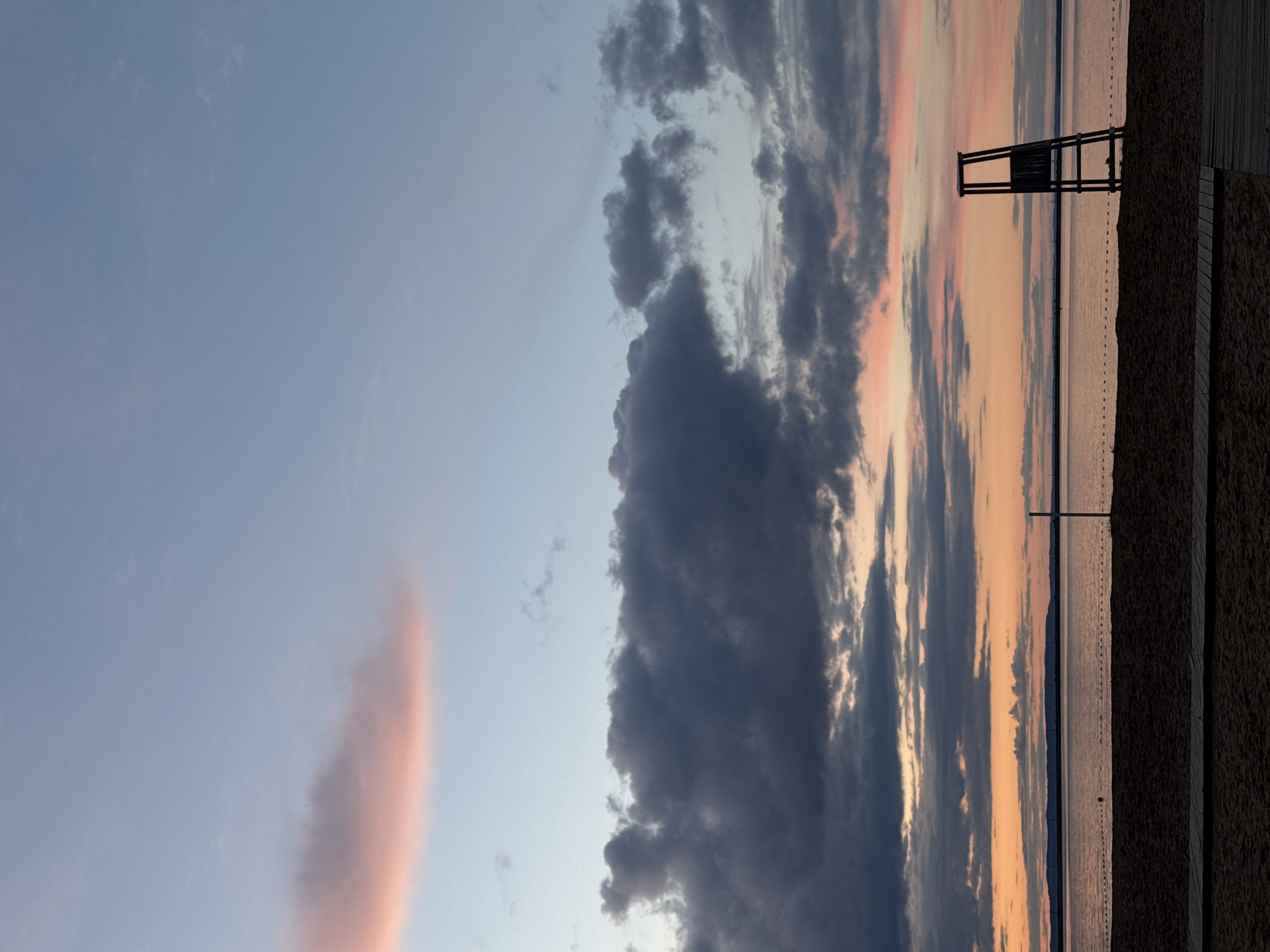 The image depicts a beach scene at sunset in Chaikovsky, Russia. A lifeguard stand is silhouetted against the sky. Clouds dominate the sky, with some areas tinted pink. The beach is visible in the foreground, with a boardwalk on the right. A body of water, likely the Kama River, stretches to the horizon. Buoys mark off a section of the water.