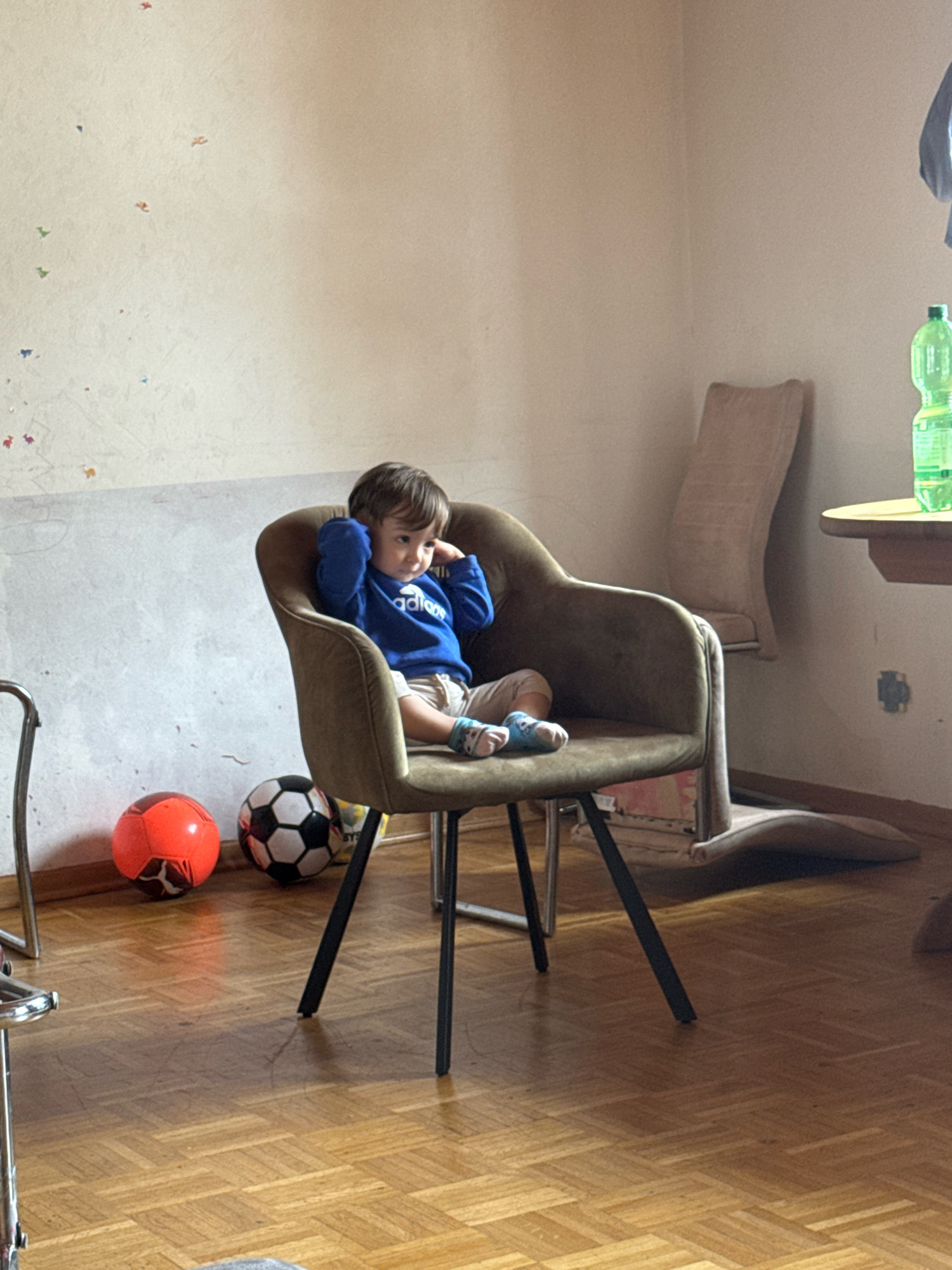 The image depicts an indoor scene in a room that appears to be a living area, possibly in Überherrn, Germany. The main subject is a young child, estimated to be between 2-3 years old, sitting in a brown armchair with their hands covering their ears. The child is wearing a blue sweatshirt with white lettering, light-colored shorts, and patterned socks.
In front of the armchair, on a wooden floor, are two soccer balls: one orange and one black and white. To the right of the armchair, a table with a light wooden top is partially visible, on which rests a green plastic bottle of liquid. In the background, a tall, light-colored chair is propped against the wall, and the wall itself has a textured, light-colored finish with faint colorful decals or drawings scattered across it. The overall lighting suggests it is daytime, with sunlight casting shadows on the floor. The room seems moderately furnished and lived-in.
