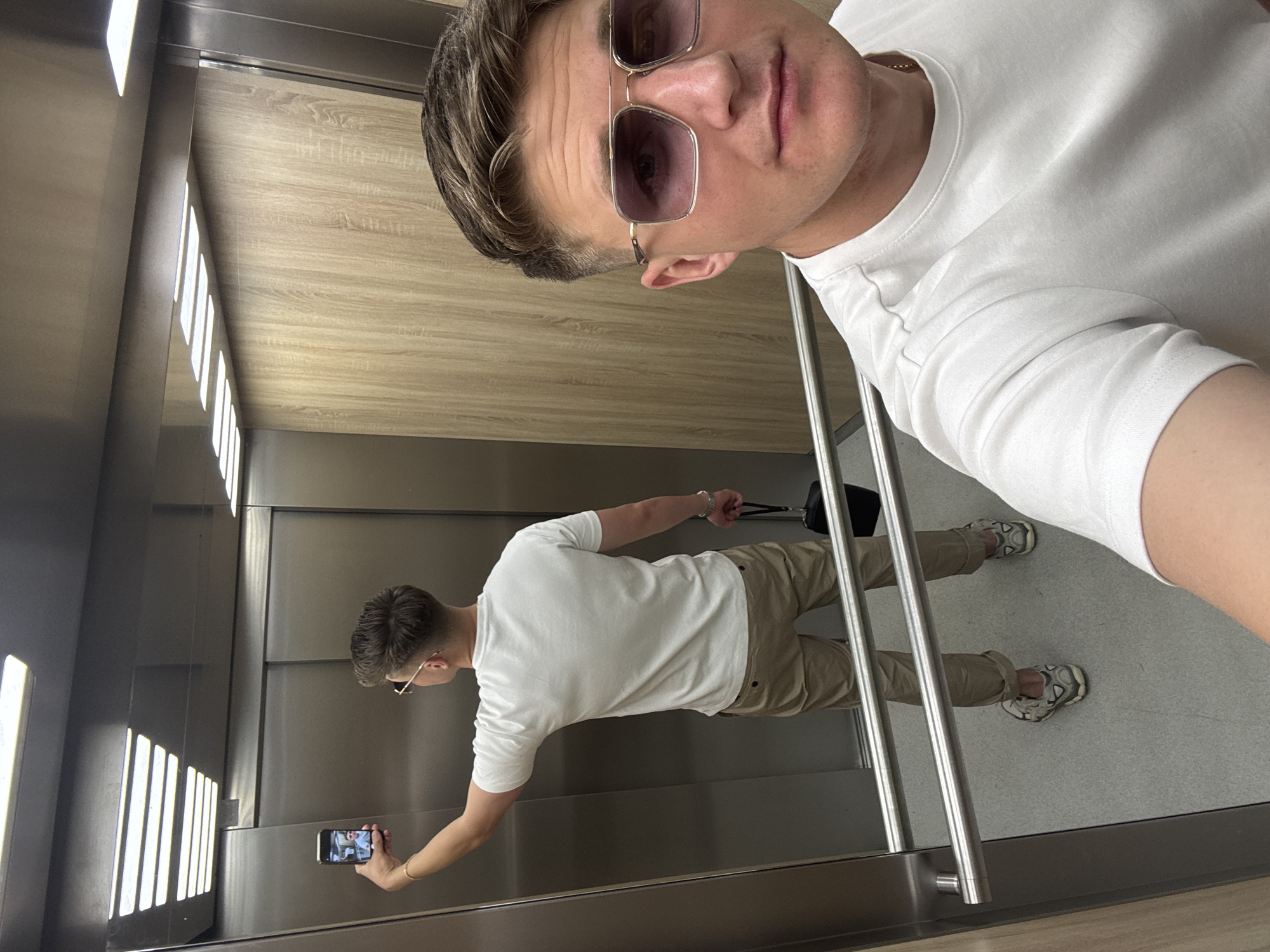 A man takes a selfie in a mirror in an elevator. The elevator's interior features metal and wood finishes. The man wears sunglasses, a white t-shirt, and khaki pants. A reflection of the man taking the photo is visible in the mirror. The location is unknown, and there is no indication of Plovdiv, Bulgaria.