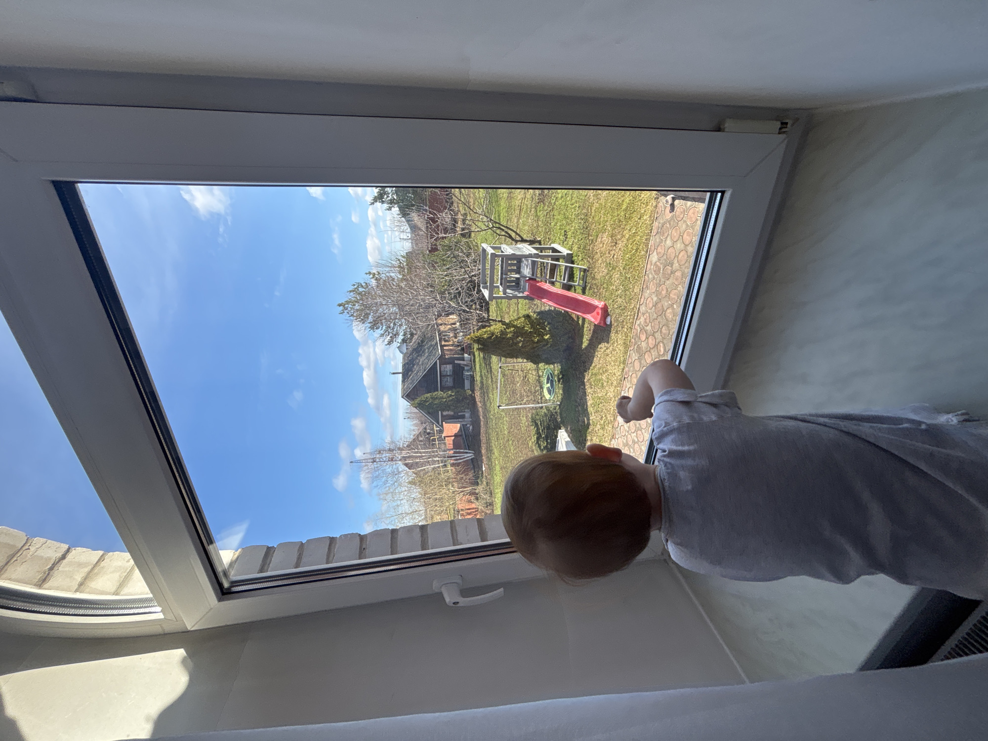 A young child in a light grey shirt stands at a window, looking out onto a yard. Outside, there is a playset with a red slide, a green lawn, trees, a building, and a blue sky with white clouds. The window frame is white. The location is Ферма, Russia.