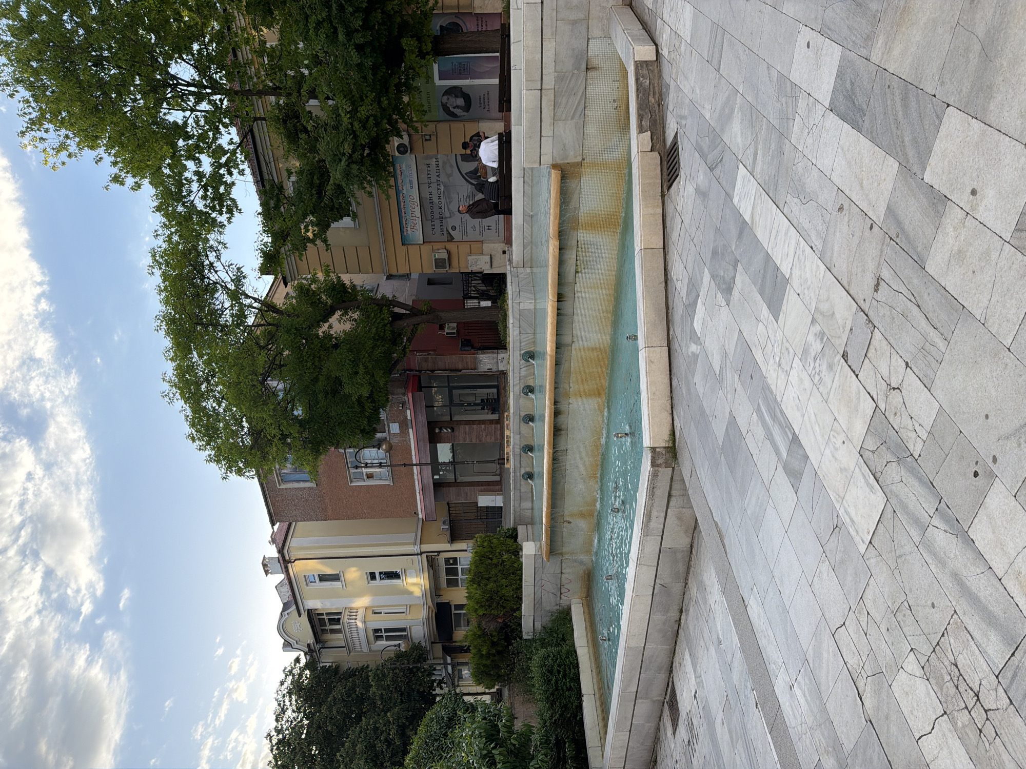 The image depicts a street scene in Burgas, Bulgaria. The foreground features a tiered fountain with a light blue water pool. Adjacent to the fountain is a tiled pavement area. Buildings line the street, and trees are present. The sky above is visible, showing a mix of blue and clouds. People are seen seated on a bench in front of one of the buildings, and there's an advertisement sign reading "Belprego" and "СЧЕТОВОДНИ УСЛУГИ БИЗНЕС КОНСУЛТАЦИИ," indicating accounting and business consultancy services. The image is oriented vertically.