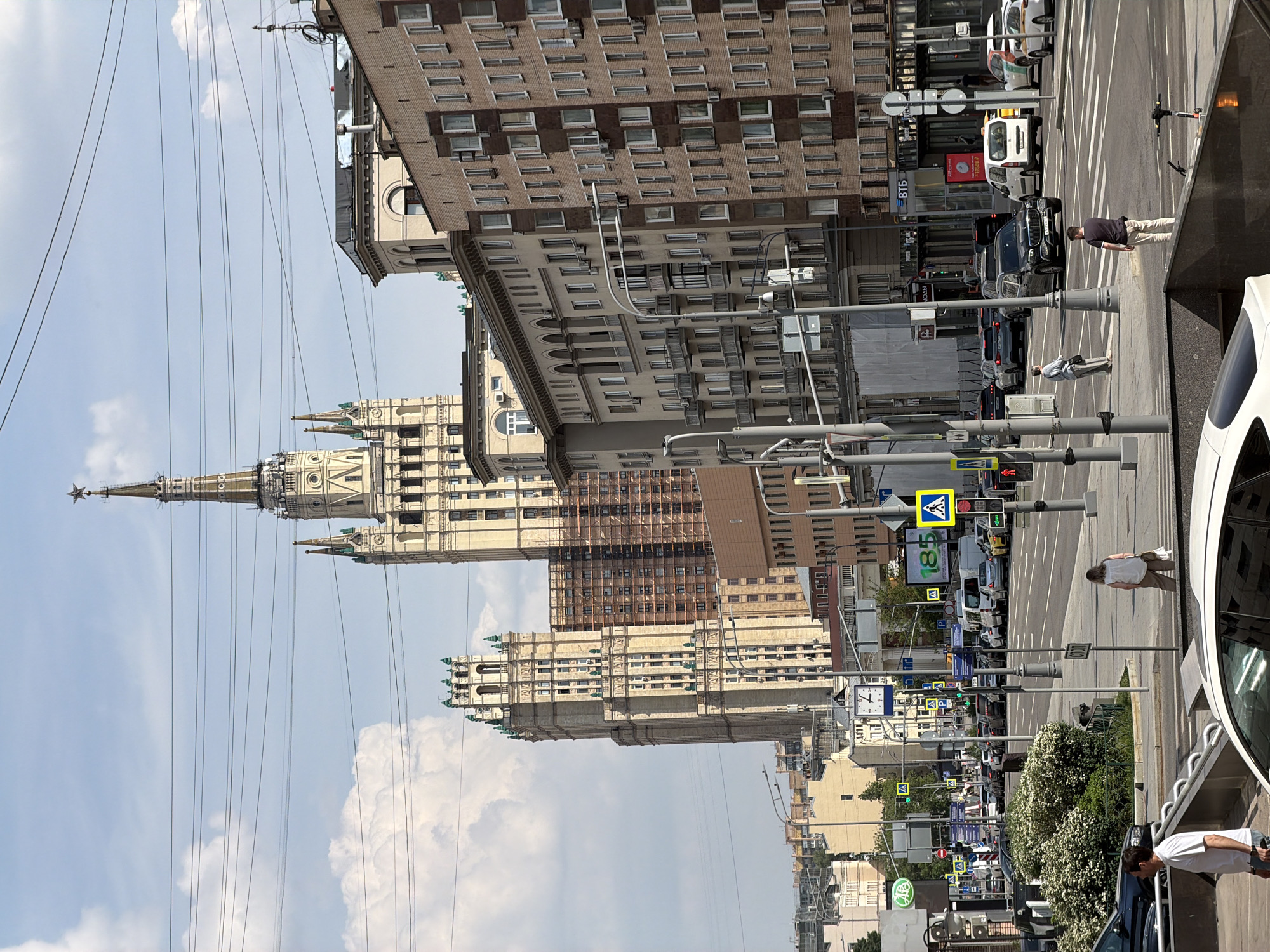 The image captures a Moscow street scene featuring prominent Stalinist architecture. In the background stands one of the Seven Sisters skyscrapers, characterized by its ornate design and central spire topped with a star. The street is lined with buildings of varying heights and architectural styles, reflecting the urban landscape of Moscow. A pedestrian crosswalk is visible, with several traffic lights and street signs posted nearby. Pedestrians can be seen crossing the street, and vehicles, including cars and vans, are present in the scene, suggesting moderate traffic. Overhanging wires run across the sky, adding to the complex urban environment. The sky is partly cloudy, casting diffused light across the scene.