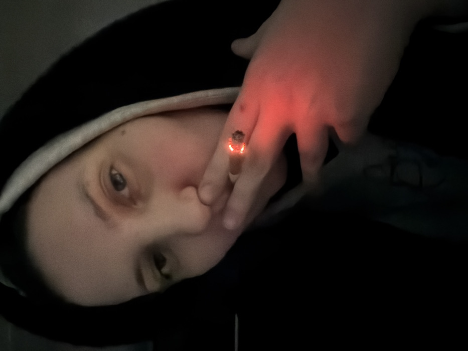 A person in a black hoodie with a white trim is the subject of this close-up shot. Their left eye is visible, and their hand is covering part of their mouth. The hand and a ring on the finger glow with a reddish-orange light, creating an eerie effect. The overall lighting is dim, adding to the image's mysterious atmosphere.