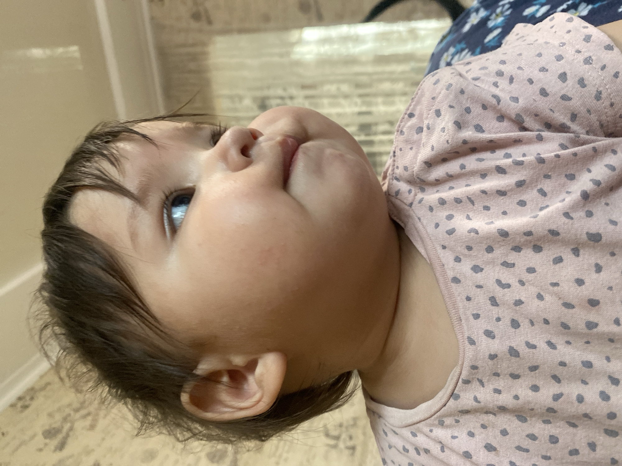 The image shows a close-up, sideways view of a baby's face and upper body. The baby has dark hair, blue eyes, and chubby cheeks. They are wearing a light pink shirt with a pattern of greyish-blue polka dots. The background is blurred but appears to be an indoor setting with a light-colored wall and floor. The baby's head is tilted upwards and to the side, and they seem to be looking at something or someone off-camera. The lighting suggests it is daytime. The overall mood is calm and intimate, capturing a candid moment with the baby. There is no visible text in the image.