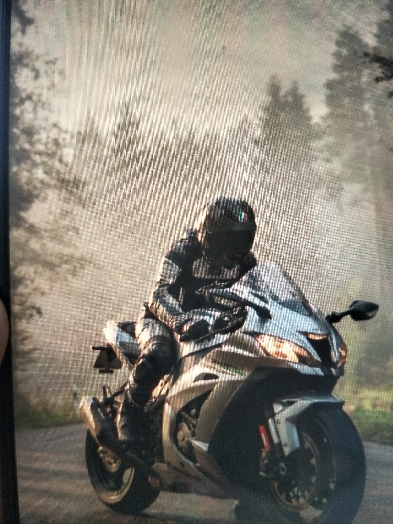 A motorcyclist wearing full protective gear, including a helmet, is riding a silver sportbike on a road. The background is a misty forest with tall trees. The scene appears to be in a natural, outdoor environment. There is no information in the image to suggest the location is the city of Payagaboh, Indonesia.