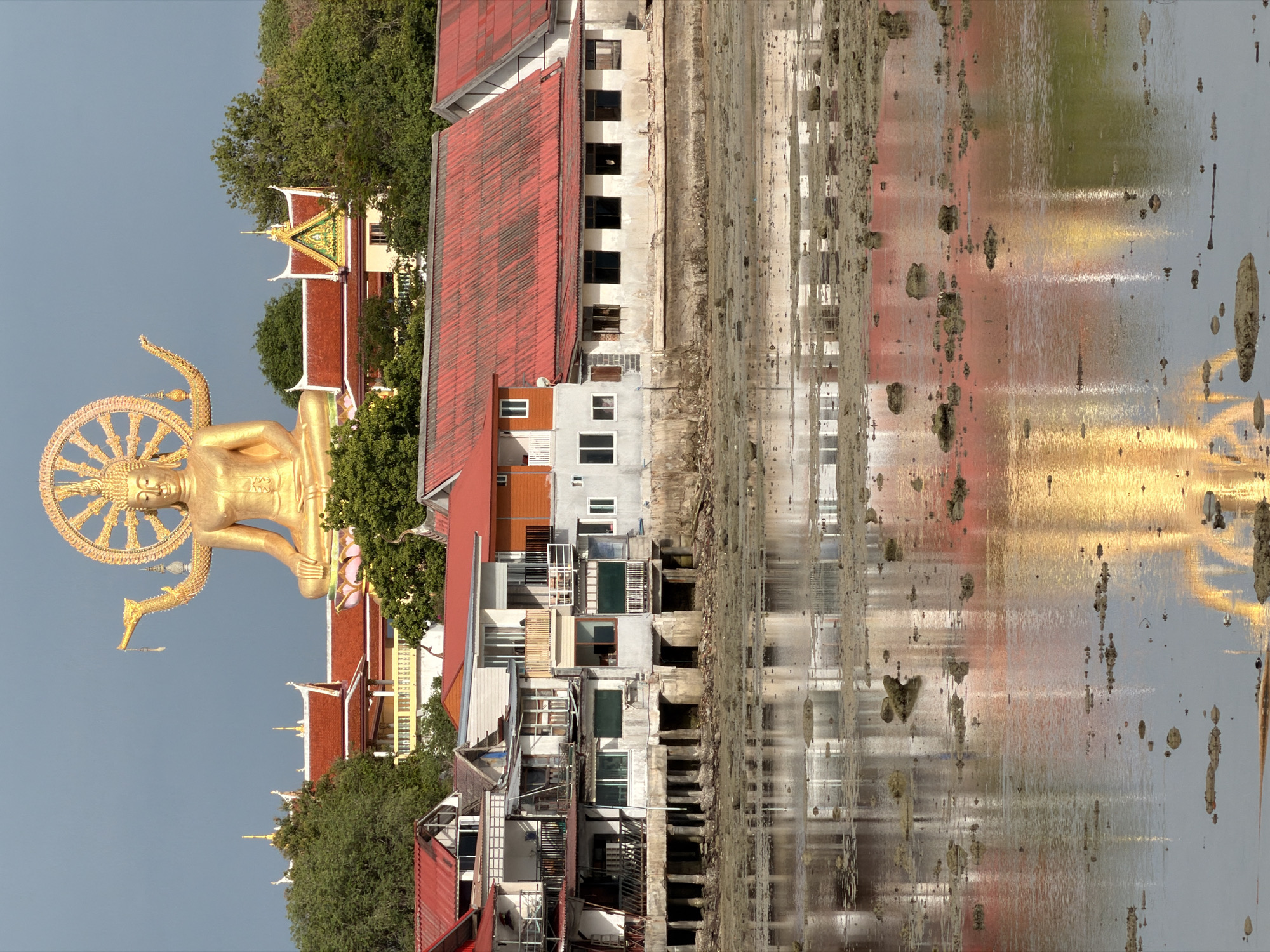 This image depicts a serene coastal scene in the city of Ko Samui District, Thailand, dominated by a massive golden Buddha statue atop a hill. The statue, seated in a meditative pose with a halo of rays, overlooks a cluster of buildings with red tiled roofs. In the foreground, a body of water, possibly a shallow bay or estuary at low tide, reflects the structures and the sky. The water's surface is partially obscured by exposed rocks and debris, suggesting a low tide condition. The architecture of the buildings closer to the water appears to be functional, possibly residential or commercial, with multiple stories and balconies. The overall atmosphere is tranquil, with the golden Buddha serving as a prominent spiritual landmark against the backdrop of the natural and built environment of Ko Samui.