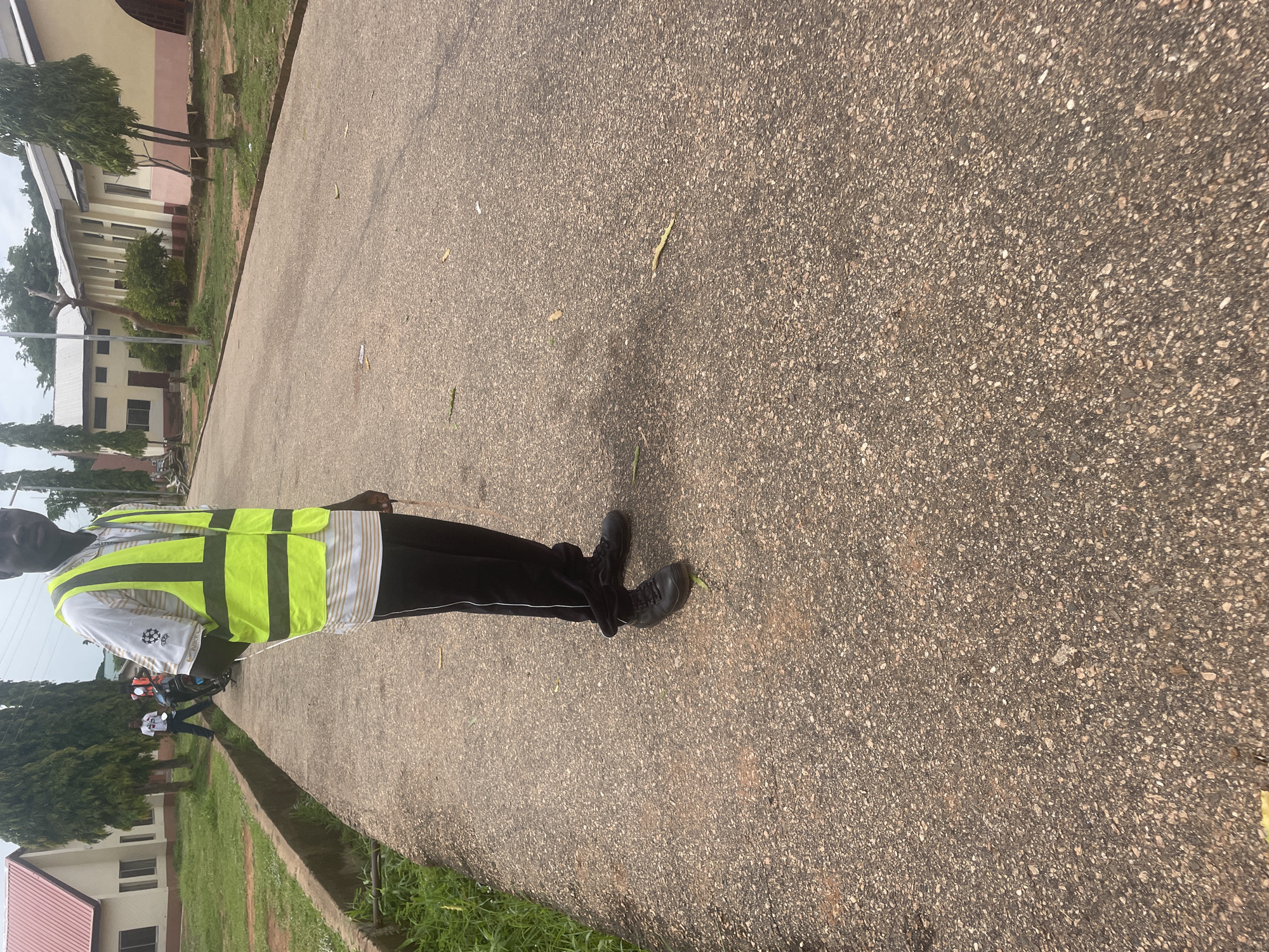 A person wearing a high-visibility vest stands on a paved road in Lokoja, Nigeria. The person is wearing a white and striped shirt, black pants, and black shoes. The vest is neon yellow with reflective stripes. In the background, there are buildings, trees, and another person on a motorcycle. The road appears to be in a campus or institutional setting, with grassy areas and a drainage ditch to the left. The sky is overcast.