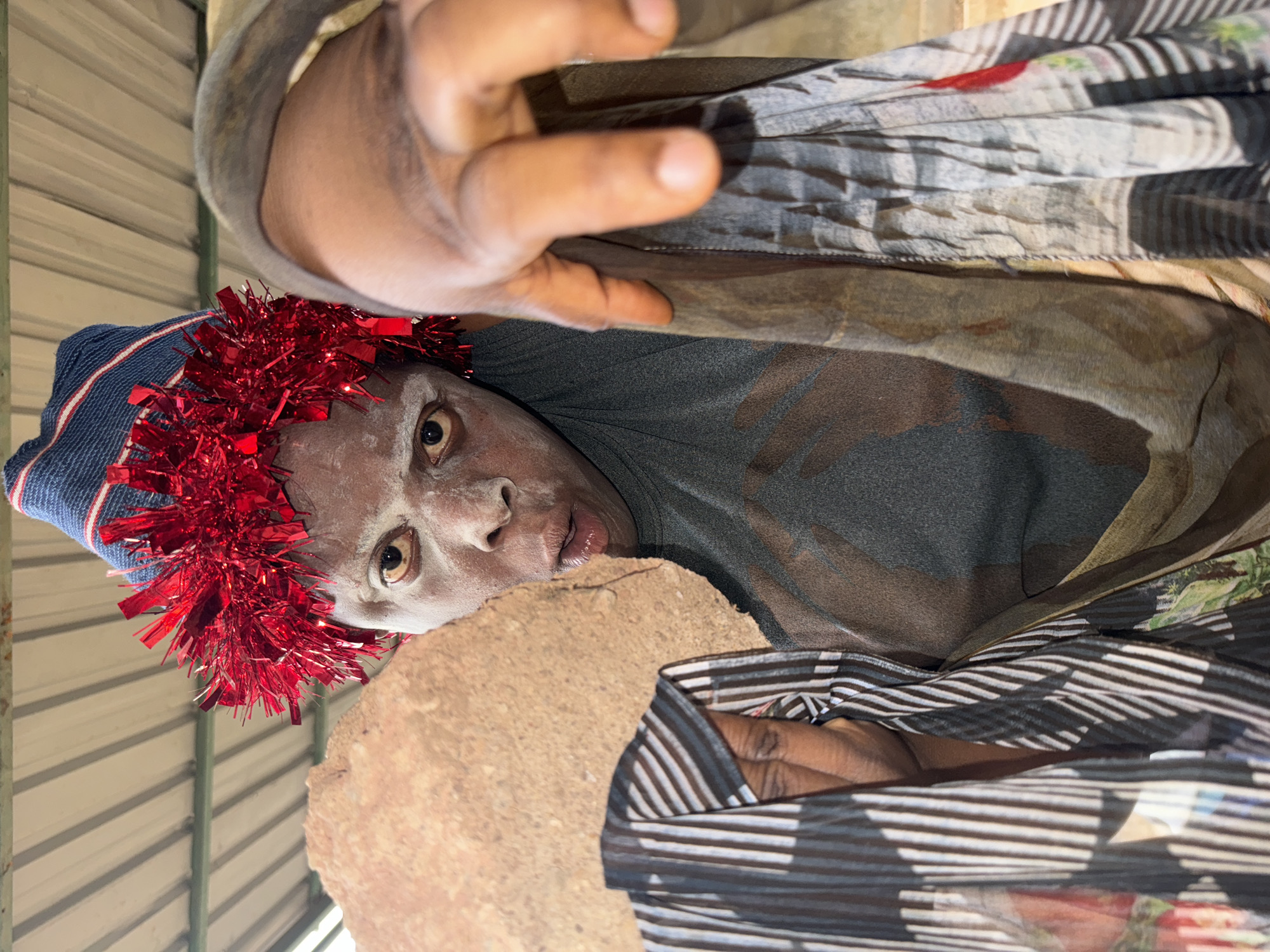 A man is shown from a low angle, possibly peering into something. He is wearing a blue and white striped hat with a red tinsel garland around it. His face is heavily covered in white powder, contrasting with his skin tone. The man has a gray shirt on, and a patterned sheer fabric is draped around him. A reddish-brown, rough-textured object is near his face. The background appears to be a corrugated metal ceiling. The location is Sabon Tasha, Nigeria.