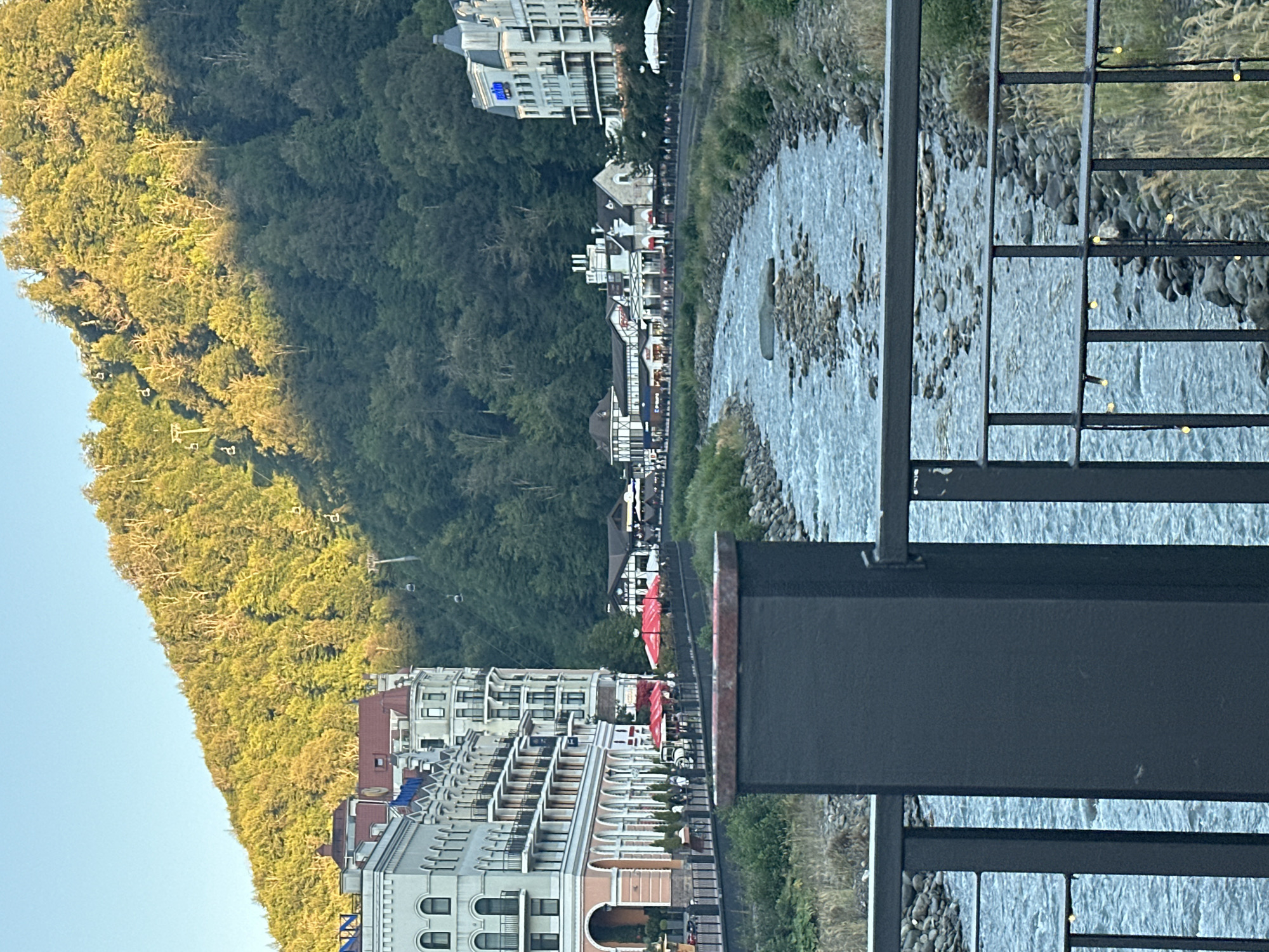 The image shows a waterside village in Estosadok, Russia. The scene is divided into two main parts by a river. On the left side, a steep, tree-covered mountain slopes downwards towards a collection of buildings. The trees on the mountain are lush green and appear to be catching the golden light of the sun. The buildings are modern in style, with multiple levels and balconies, some featuring reddish-brown roofs. On the right side of the river, more buildings are visible, including some with a more traditional alpine architectural style, featuring dark timber accents and white walls. Red umbrellas are set up outside some of these buildings, suggesting outdoor seating for cafes or restaurants. The river itself is wide and appears shallow, with a rocky bed visible through the clear, flowing water. The foreground is dominated by a dark metal railing, suggesting the photo was taken from a balcony or walkway overlooking the scene. The lighting indicates it is daytime, likely late afternoon given the warm sunlight on the mountain. The weather appears clear and pleasant. There is no visible activity or specific event taking place, but the setting suggests a bustling tourist destination. Some text is visible on the buildings, including what appears to be a blue logo with white lettering on one of the larger buildings.