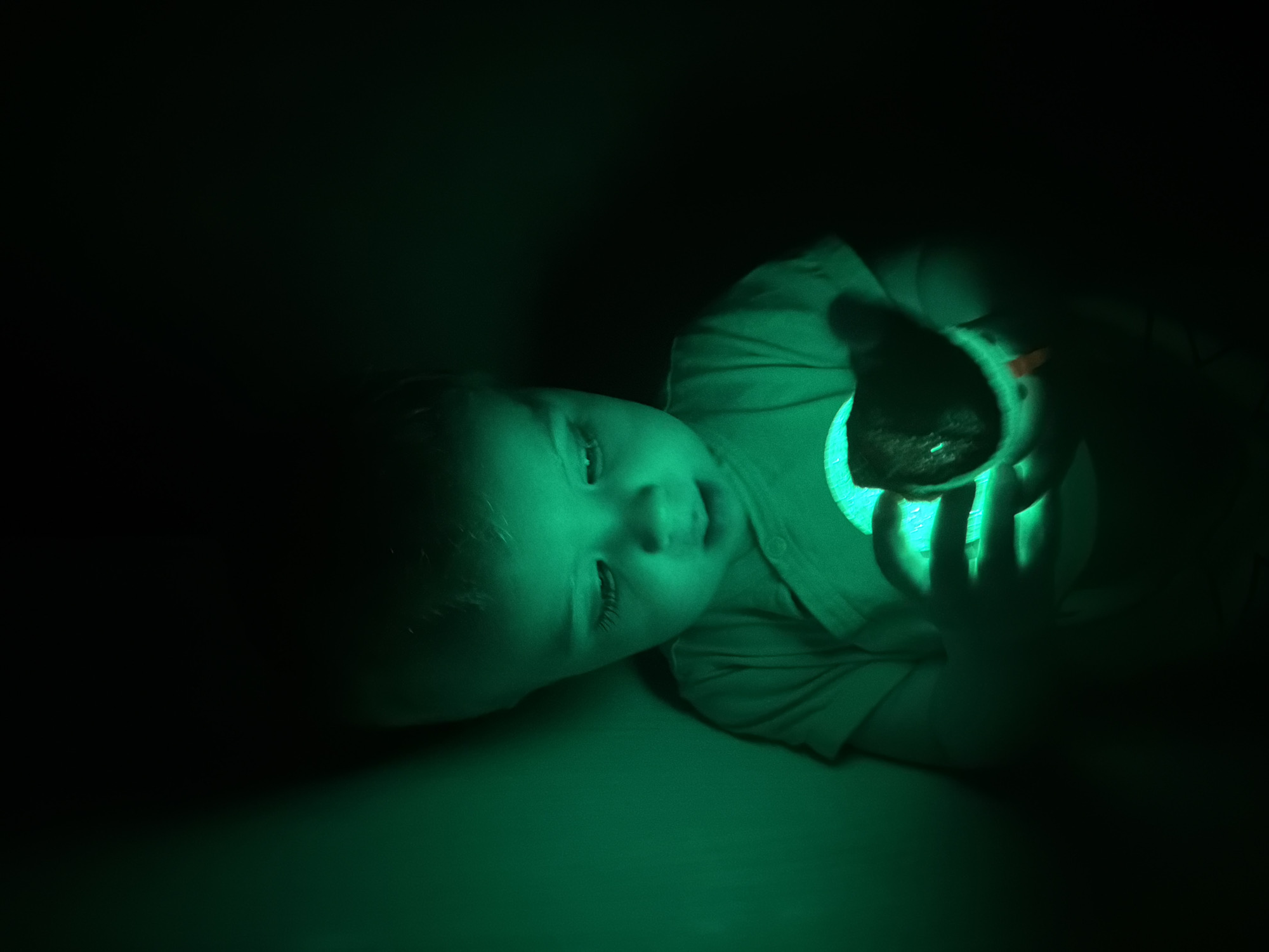 A young child is lying down in a dark environment, illuminated by a green light emanating from an object they are holding. The child is fair-skinned with light hair, looking up with a slight smile. They are wearing a light-colored shirt. The object in their hands appears to be a plush toy with a light source inside. The scene is predominantly dark, with the green light source casting shadows and highlighting the child's face and hands. There are no indications of the location being Votkinsk, Russia.
