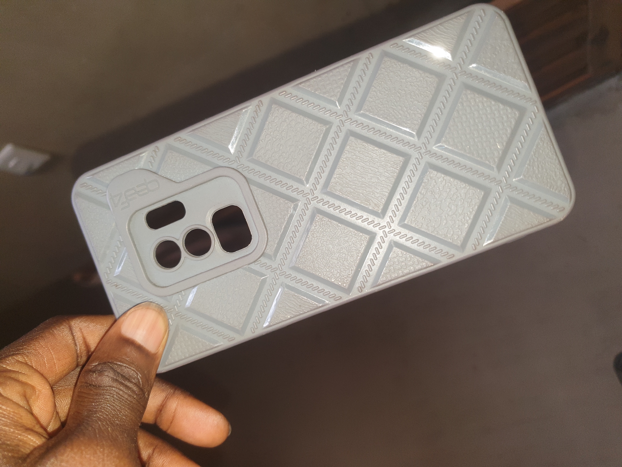 In an indoor setting with a blurred brown background, a person's hand with dark skin and short fingernails holds a light gray phone case. The phone case features a raised, textured diamond pattern across its surface, with a rectangular camera housing at the top. The camera housing has three circular cutouts. The word "CESO" is embossed in a subtle, light gray font on the top left of the camera housing. The phone case is tilted slightly, with the bottom right corner closer to the viewer.