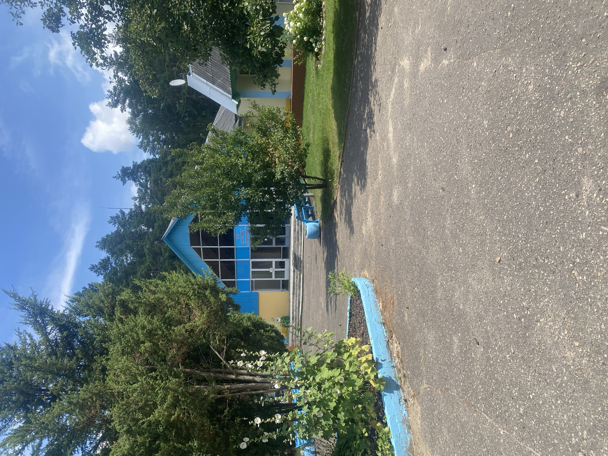 A wide shot shows a bright blue building with a large glass entrance, framed by lush green trees and a clear blue sky with scattered clouds. The building is situated in a rural setting with a paved walkway leading to its entrance. The walkway is bordered by a low, blue-painted wall and a flower bed containing tall white flowers. To the right of the blue building, a smaller yellow building with a grey, sloped roof is visible. The scene appears to be a public space, possibly a community center or a school, given the signage on the blue building which reads "ЗАЛ МАССОВЫХ МЕРОПРИЯТИЙ" (Hall for mass events). The overall impression is one of a well-maintained, sunny outdoor area in a peaceful, natural environment. This location is identified as being in Ilyanskiy selskiy Sovet, Belarus. There are no people or animals visible in the image, and no activities are taking place.