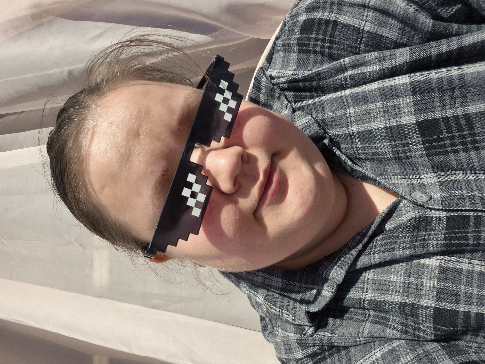 A woman with light skin and light brown hair pulled back is wearing "deal with it" pixelated sunglasses and a grey plaid shirt. She is lying down, possibly outside, with a white or light-colored surface visible in the background. The location is not determinable as Chaikovsky, Russia.