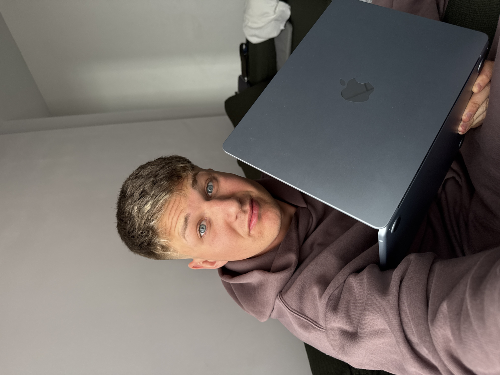 A young Caucasian male with short blonde hair and blue eyes is holding a closed grey laptop in a portrait-style shot. He is wearing a mauve-colored hoodie. The background includes a cream-colored wall and a dark green couch with a white cloth on it. The location is not specified but has nothing to do with the city of Plovdiv, Bulgaria. The man is looking directly at the camera.