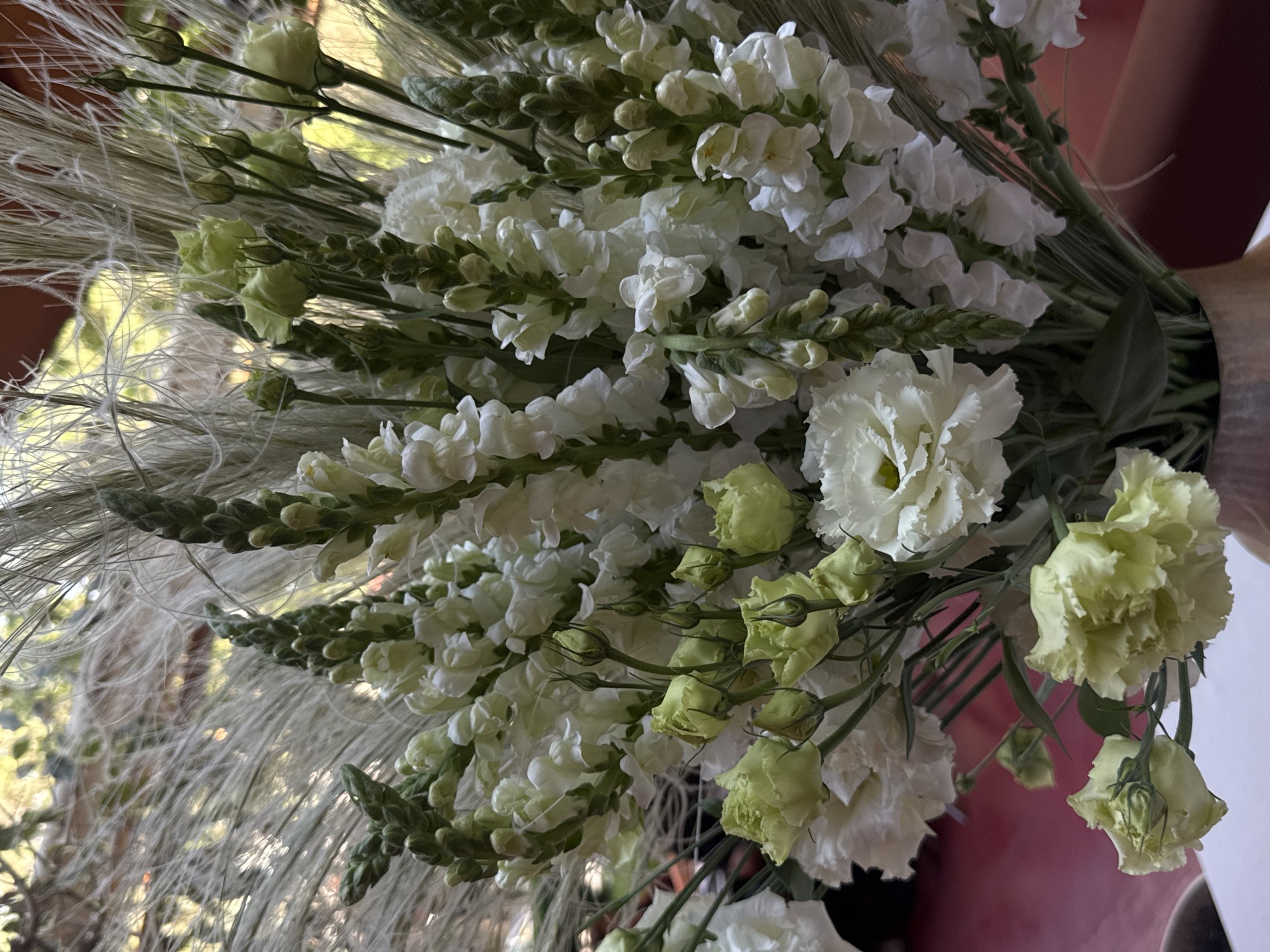 This is a close-up, eye-level shot of a floral arrangement in a vase. The arrangement features a mix of white snapdragons and light yellow roses, along with some dried grass elements. The flowers are tightly packed and occupy most of the frame, with some of the vase visible at the bottom. The background is blurred, with hints of green foliage and a reddish-brown surface. There are no people or discernable actions taking place. The image does not contain any visual cues that would indicate its location in Yerevan, Armenia.
