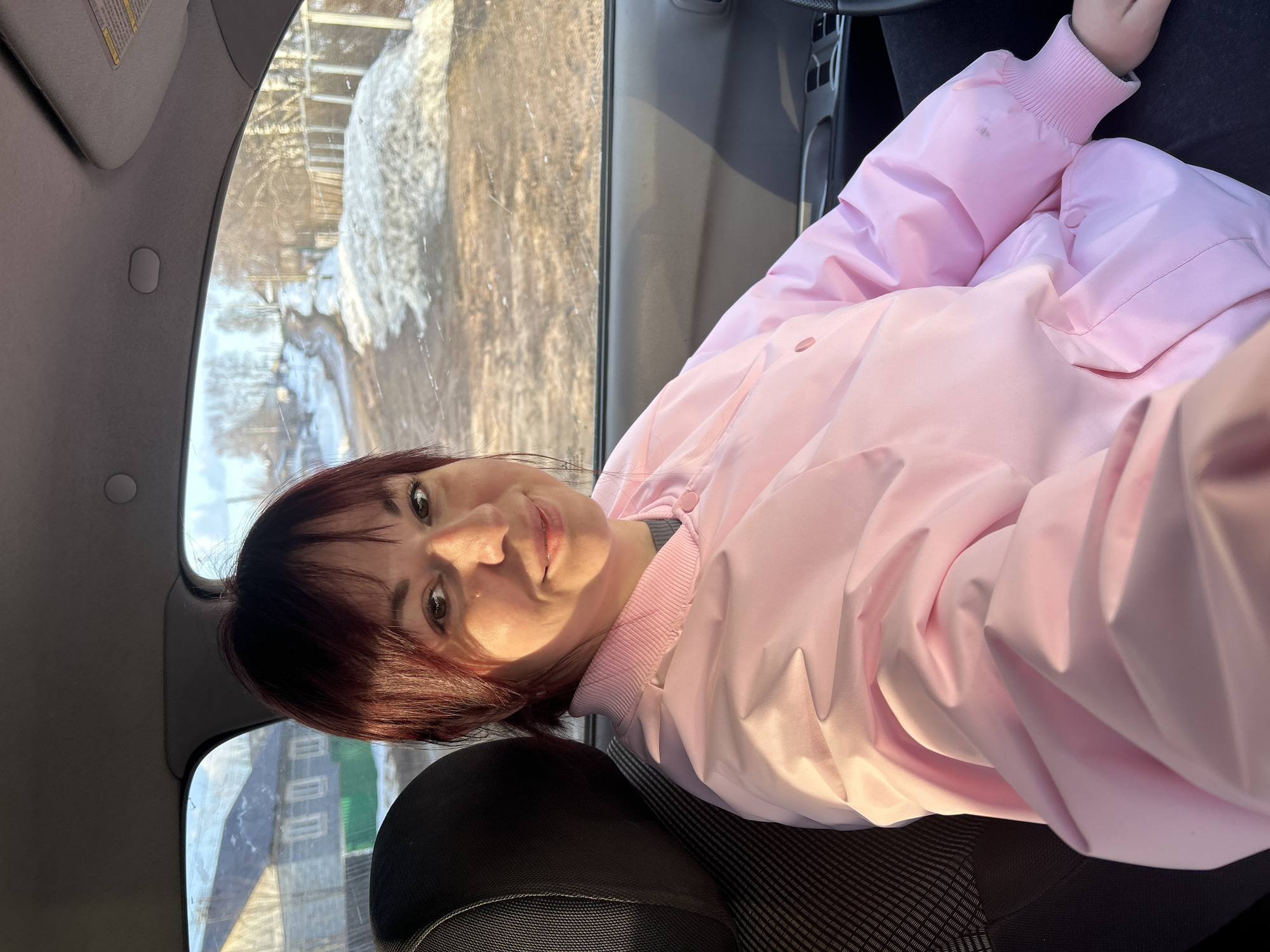 A woman with dark hair and a light pink jacket is sitting in the driver's seat of a car. The car is parked in Фоки, Russia, and through the window, there is snow and a building that is green and white. The woman is looking at the camera.