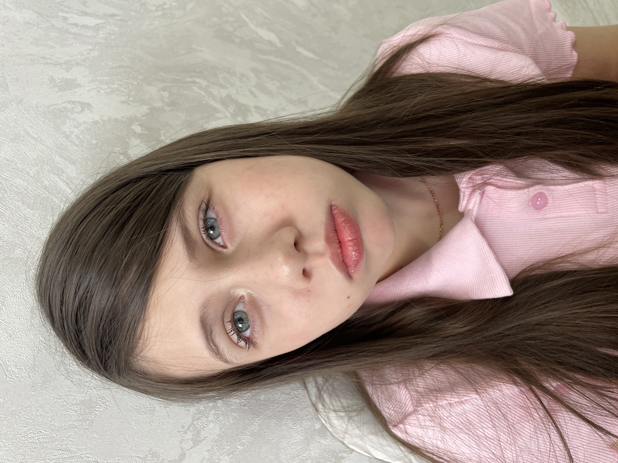A fair-skinned young woman with long, dark brown hair and light blue eyes is the subject of this close-up shot. She is wearing a pink collared shirt with a button visible at the collar. Her lips are a soft pink color with a glossy sheen. The background is a textured, off-white surface.
There is no information in the image about the city of Izhevsk, Russia.