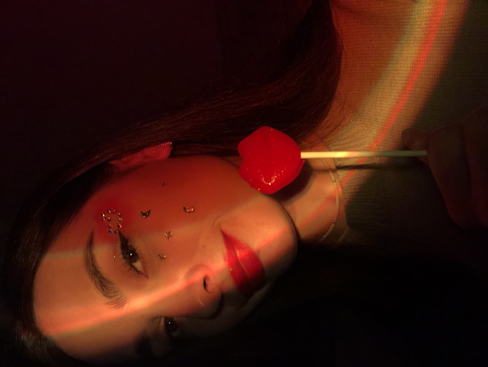 A woman with light skin and dark hair is pictured lying on her side in a dark setting, illuminated by a red light streak across her face. She has red lipstick and small decorative stickers on her forehead near her eyebrow. She is holding a red heart-shaped lollipop on a stick near her cheek. She is wearing a light-colored knitted sweater. The background is mostly dark with a muted light pattern on the upper right. There is no indication of this photo being taken in Noviy, Russia.