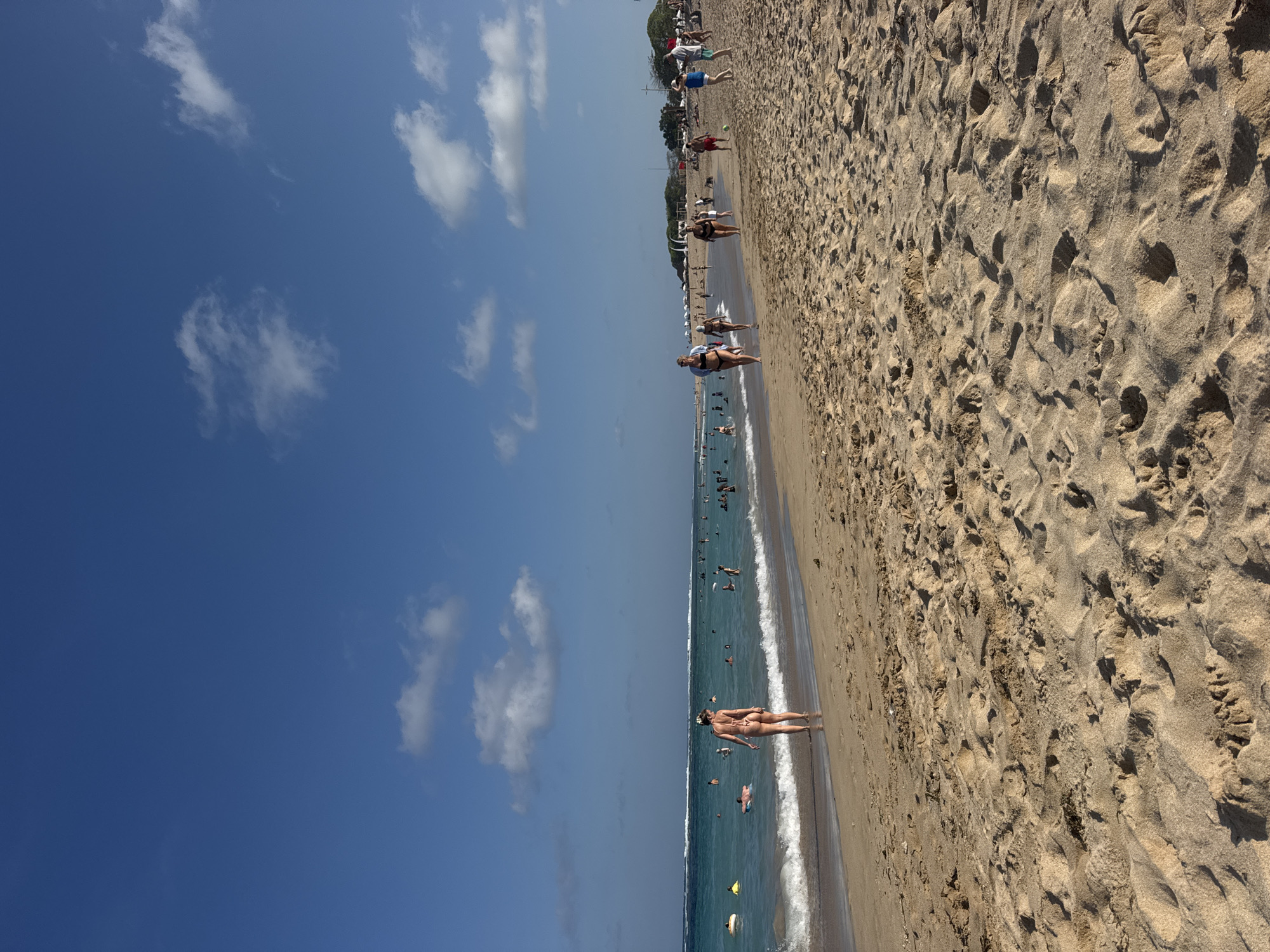 The image depicts a wide, sandy beach in Kuta Selatan, Indonesia. The sky is a vibrant blue with scattered white clouds. The ocean is calm and turquoise, with gentle waves breaking at the shore. Several people are visible in the water, some swimming, others relaxing on inflatable tubes. On the beach, many individuals are walking or standing, enjoying the sunny weather. The sand is a warm, light brown, showing footprints and indentations. In the background, there are some trees and structures, suggesting the presence of resorts or beachside facilities. The scene captures a typical day at a popular beach destination.