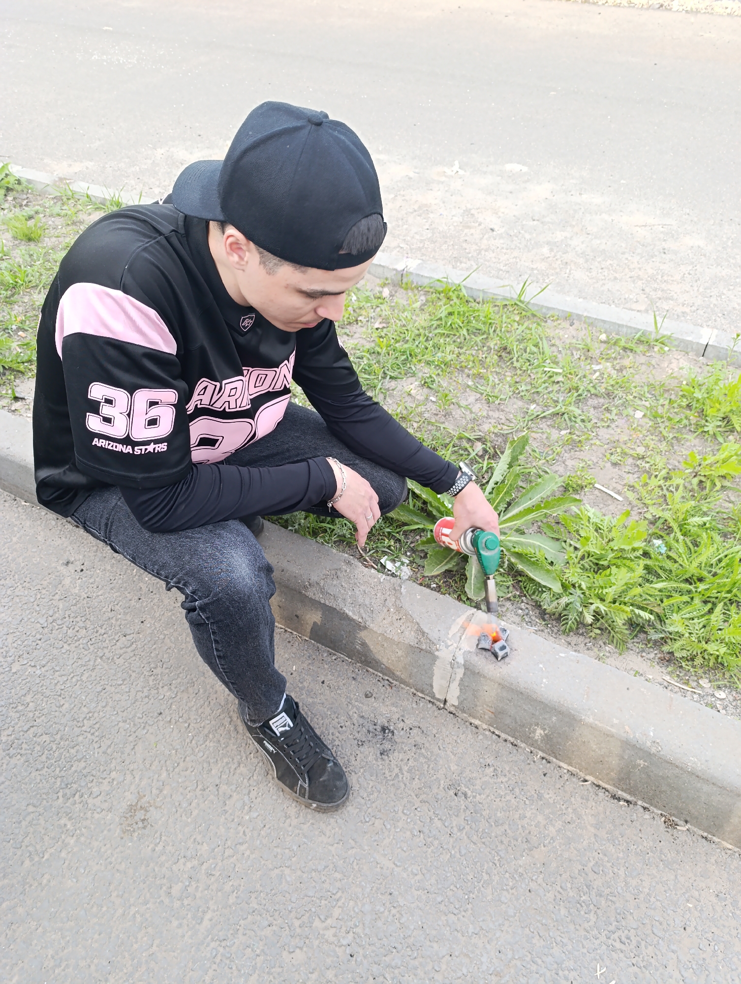 A young man is sitting on a curb next to a street in Unknown, Unknown. He is wearing a black baseball cap, a black and pink "Arizona Stars 36" jersey, black jeans, and black Puma sneakers. He is holding a small torch and appears to be burning something on the curb. The area next to the curb is covered in grass and weeds.