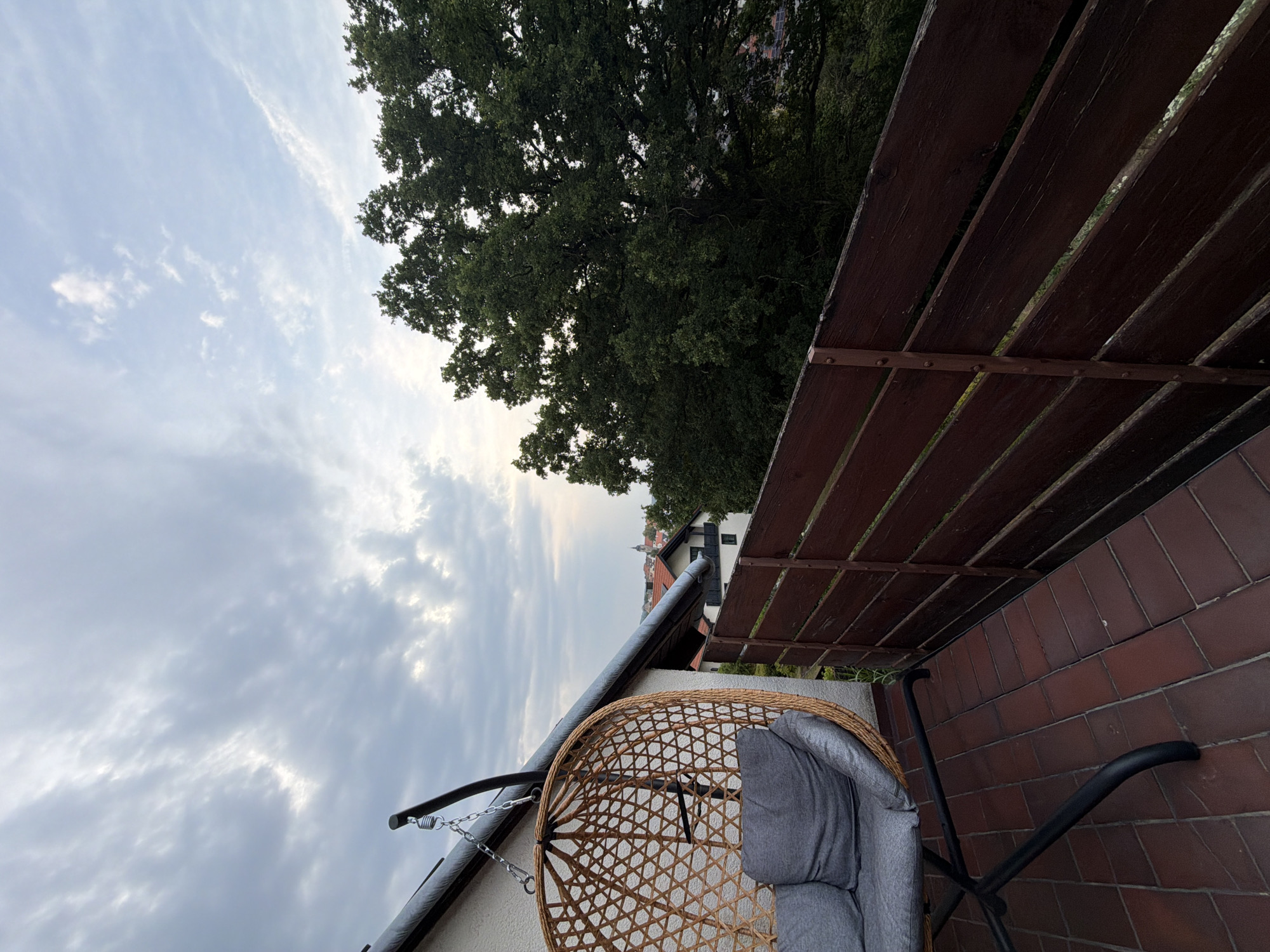 The image shows a close-up view of a balcony in Creußen, Germany. In the foreground, a wicker hanging chair with gray cushions is suspended by chains from an overhang. The balcony floor is tiled with dark red bricks. A dark brown wooden fence runs along the right side of the balcony. Beyond the fence, a large green tree with dense foliage dominates the mid-ground. In the background, a few buildings with red roofs are visible, and a cloudy sky with patches of blue is overhead. The overall scene depicts a tranquil outdoor space with a view of a town and nature.