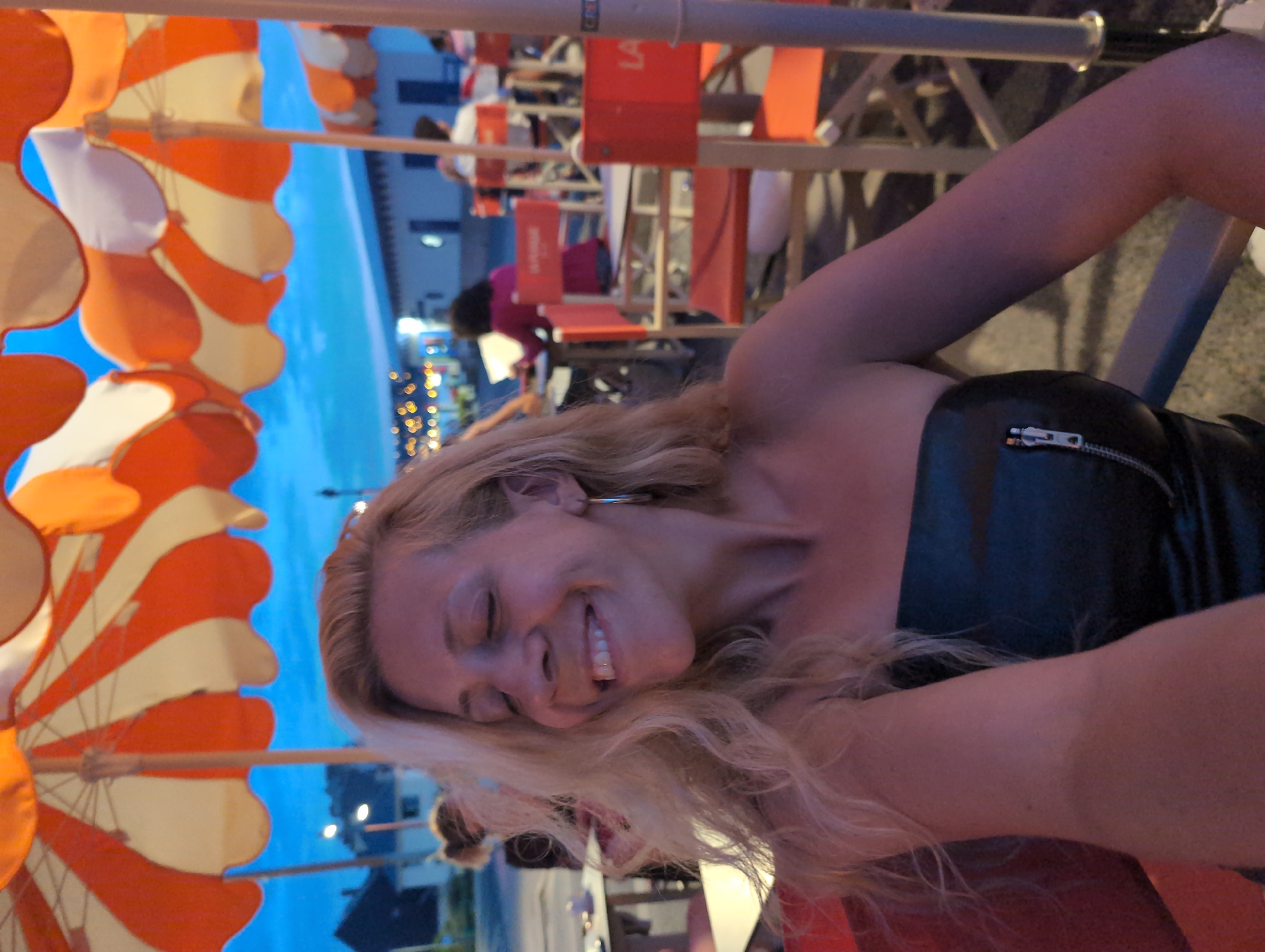 The image captures a woman with blonde, wavy hair smiling with her eyes closed, seated at an outdoor cafe. She is wearing a strapless black garment with a zipper. In the background, there are orange and white striped umbrellas, tables, and chairs. Other patrons are visible in the distance, some seated at tables. The setting appears to be a public space, possibly a promenade or street in Mers-les-Bains, France, given the distinctive architecture and ambiance suggestive of a coastal town. The sky is a deep blue, indicating it is evening or dusk. The overall scene conveys a relaxed and social atmosphere.