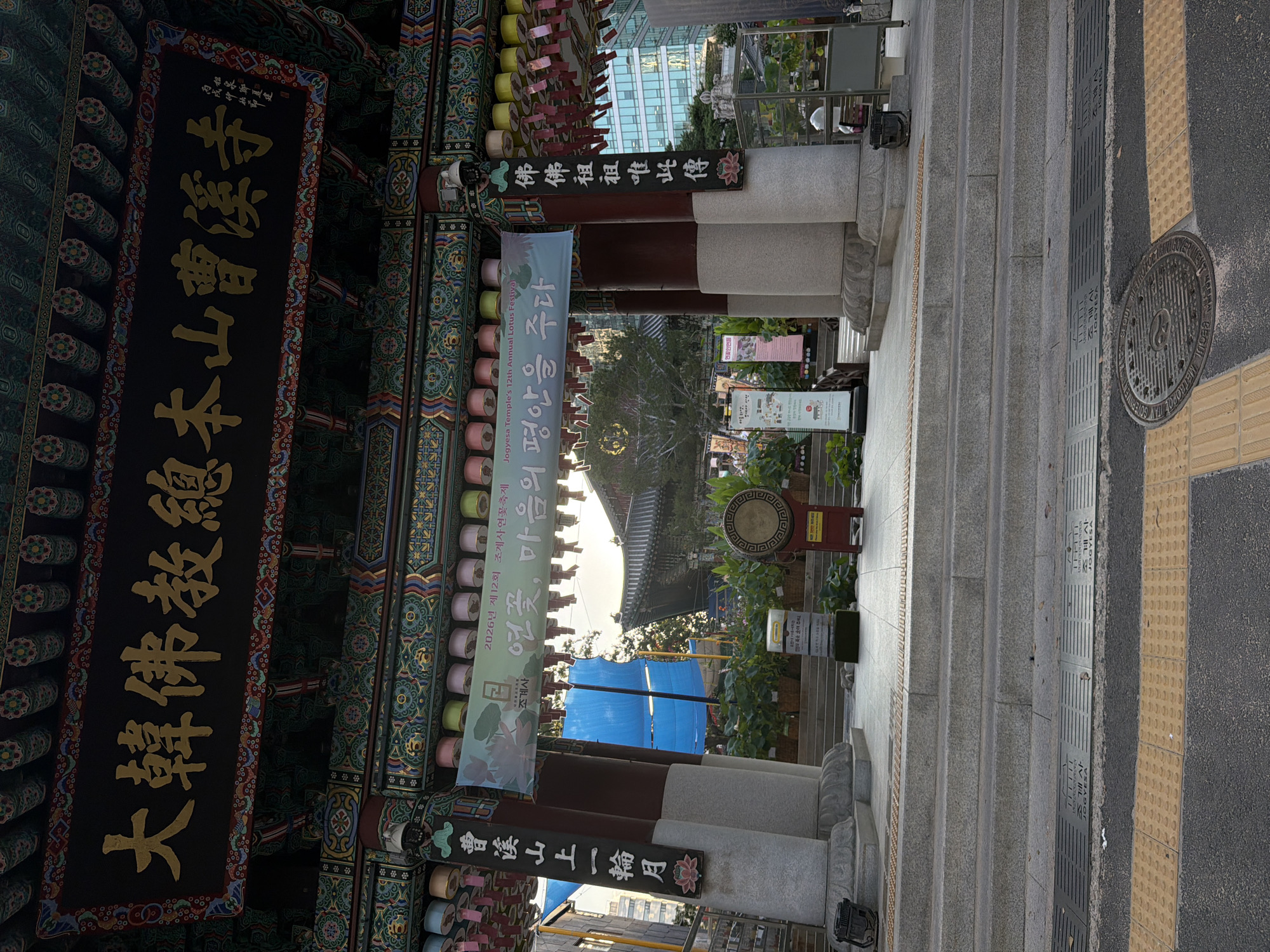The image shows the entrance to Jogyesa Temple, a prominent Buddhist temple located in the heart of Seoul, South Korea. The temple's ornate gateway features traditional Korean architectural elements, including colorful painted eaves and hanging lanterns. A large banner displaying Korean text and images of lotus flowers, along with English text reading "Jogyesa Temple's 12th Annual Lotus Festival," is prominently displayed across the entrance.
In front of the gateway, a set of wide stone stairs leads up to the temple grounds. To the right of the stairs, a large, circular drum mounted on a red stand is visible, likely used for religious ceremonies. The area is paved with stone tiles, and a yellow tactile paving strip runs along the edge of the walkway. A manhole cover with the inscription "SEOUL METROPOLITAN GOVERNMENT" is visible on the road in the foreground. The background reveals the modern cityscape of Seoul, with tall buildings contrasting with the traditional temple architecture. The lighting suggests it is daytime, with soft sunlight filtering through the trees and structures.
