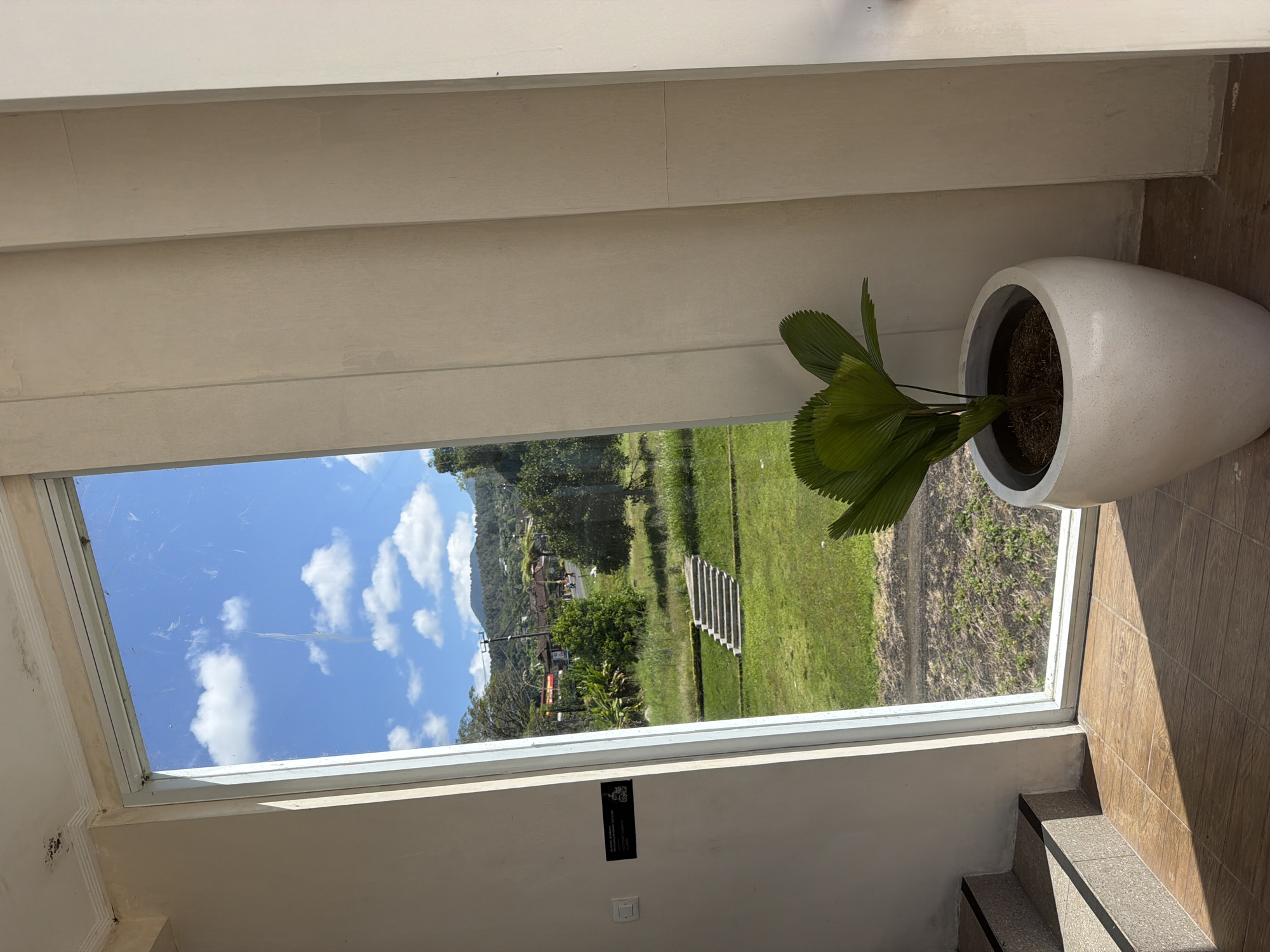 An outdoor scene through a large window reveals a lush green landscape in Baturiti, Indonesia, under a bright blue sky dotted with white clouds. In the foreground, inside the building, a large white pot contains a leafy green plant. To the left, a set of stairs with speckled treads leads upwards. The floor is tiled with a wood-grain pattern. A sign with text and graphics is affixed to the interior wall to the left of the window. The exterior view shows a grassy lawn with a set of concrete steps, trees, and buildings in the distance, with rolling hills and mountains forming the backdrop.