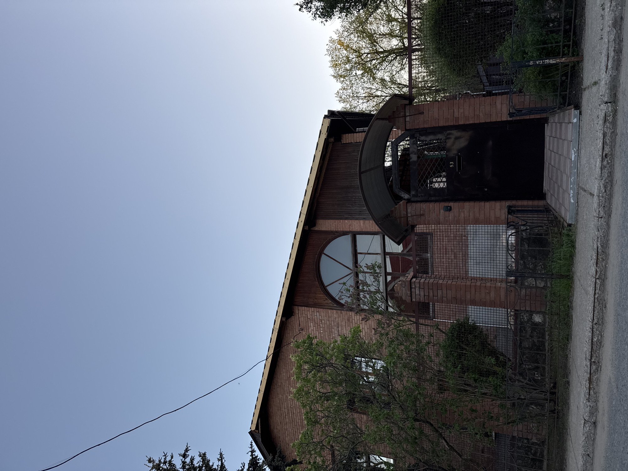 The image shows a brick building with a large arched window and a black door with a cage-like covering over the entrance. The building is surrounded by trees and bushes, and a sidewalk runs along the side of the building. There is a metal fence partially visible. The sky is a pale blue. The location appears to be in a residential area of an unknown city.