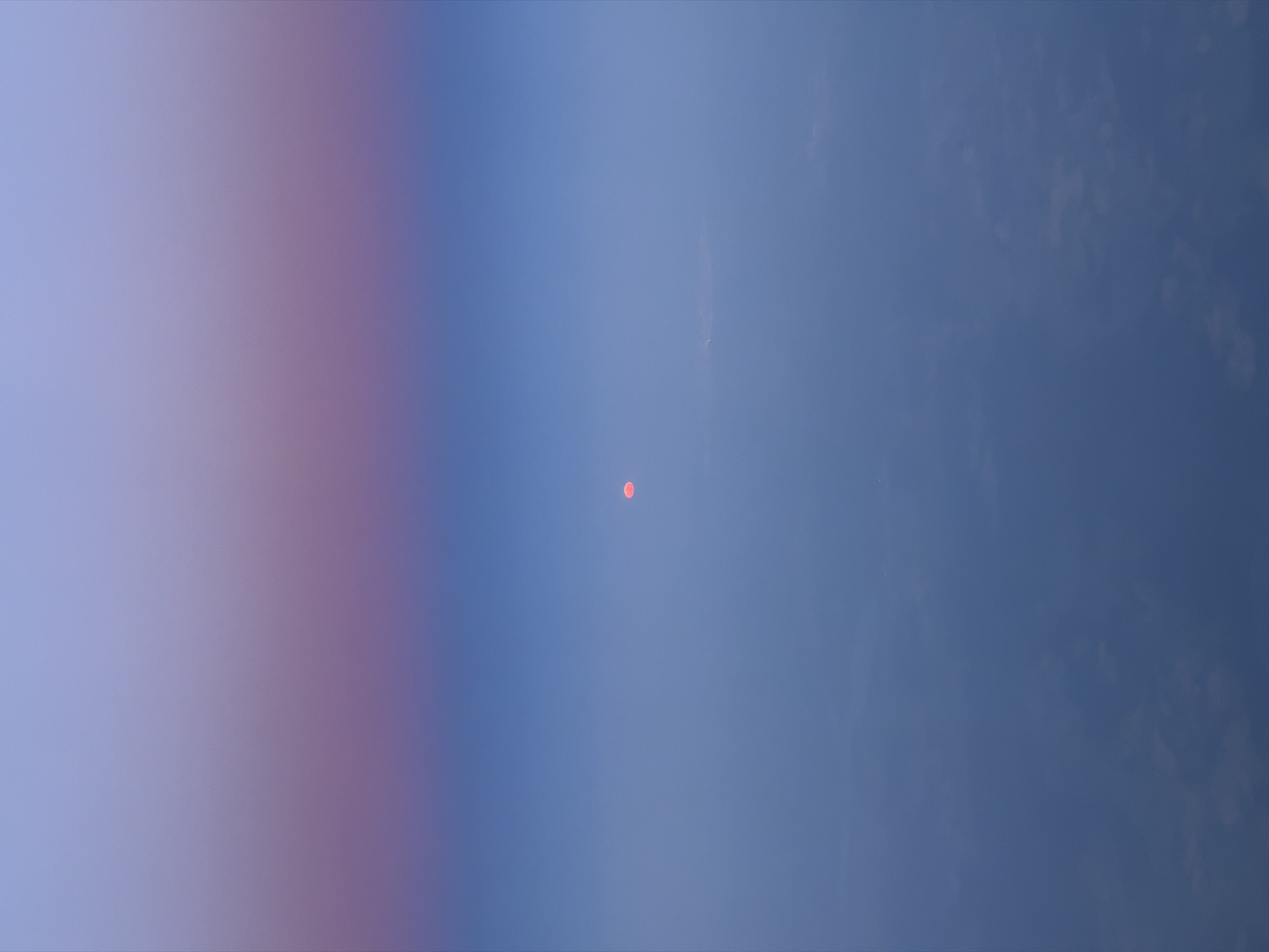 The image shows a blue sky with varying shades. On the left, the color fades from light purple to light blue. Towards the right, the blue becomes more intense. In the center of the frame is a small, circular, orange object. The sky has some faint cloud-like textures. The location is Unknown, Unknown.