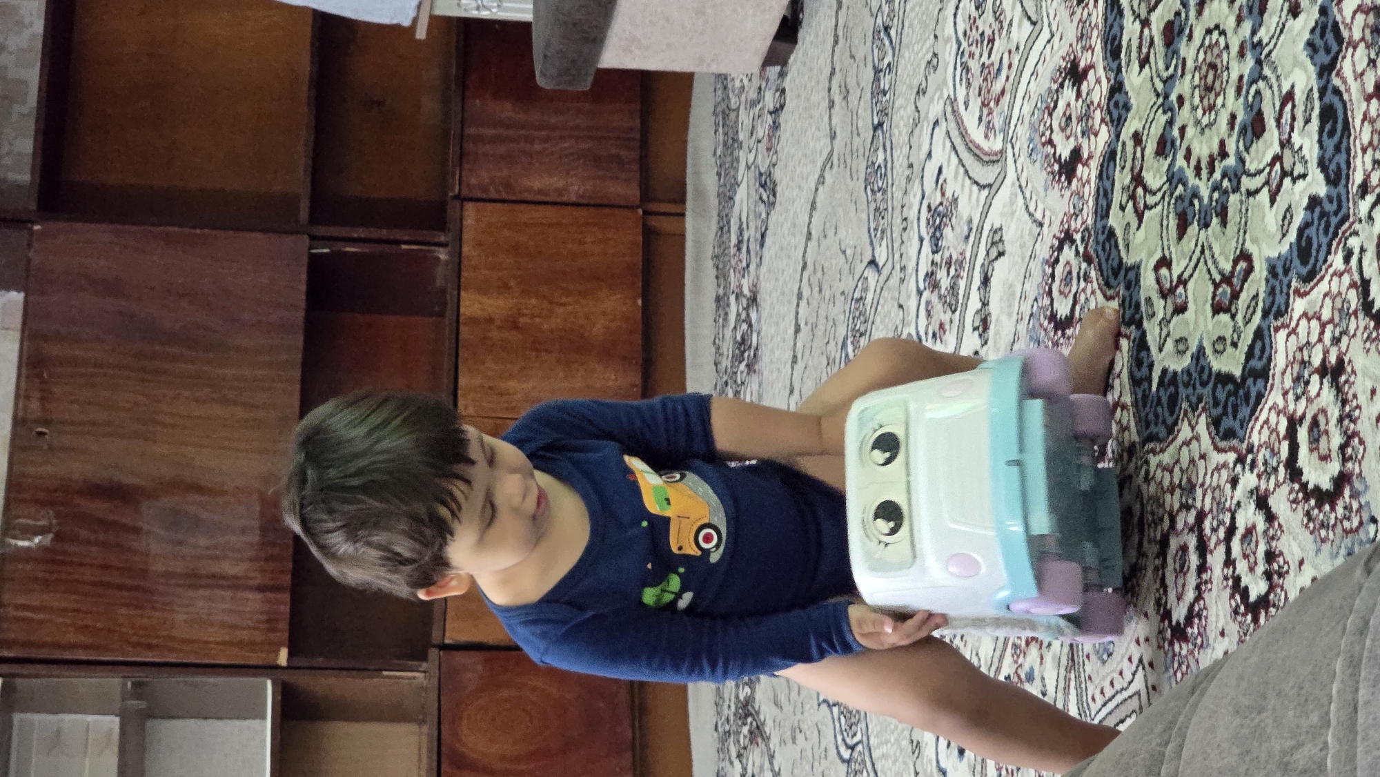 A young child, appearing to be between two and three years old, is seated on a toy vehicle indoors. The child is wearing a dark blue long-sleeved bodysuit with a graphic of a yellow truck with green accents. The child's legs are visible, extending downwards from the toy. The toy vehicle is white and light blue with large, cartoonish eyes and pink wheels. The child is looking down at the toy, and their hands are positioned on either side of it. The setting is an indoor environment with a patterned carpet covering the floor. In the background, there is a large, dark wooden cabinet with multiple shelves. This scene is located in the city of Yakkabog, Uzbekistan.