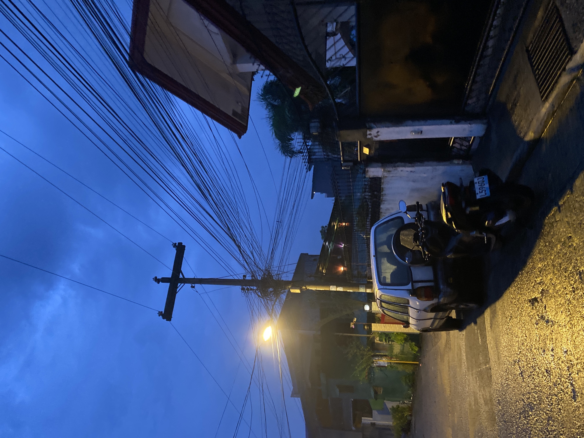 This media file captures a street scene in Davao City, Philippines, during twilight. The sky is a deep, dusky blue, suggesting the early evening or late afternoon. A prominent wooden utility pole with numerous tangled electrical wires dominates the left side of the frame, extending upwards against the darkening sky. A street light attached to the pole is illuminated, casting a warm glow on the wet asphalt road below, indicating that it may have recently rained or the street is being cleaned.
Parked on the right side of the road are a white car and a dark motorcycle. The car appears to be a hatchback, and the motorcycle is positioned behind it. The license plate of the motorcycle is partially visible and displays the text "L596QJ". The wet road surface reflects the yellow light from the streetlamp and other ambient lights from the buildings, creating a glistening effect.
The buildings lining the street are partially visible, with architectural elements suggesting a residential or mixed-use area. One building in the foreground has a dark overhang with a red trim. Other buildings further back show glimpses of windows and lights. The overall mood of the image is quiet and atmospheric, conveying the transition from day to night in an urban environment. The scene appears tranquil, with no people visible, emphasizing the stillness of the moment.
