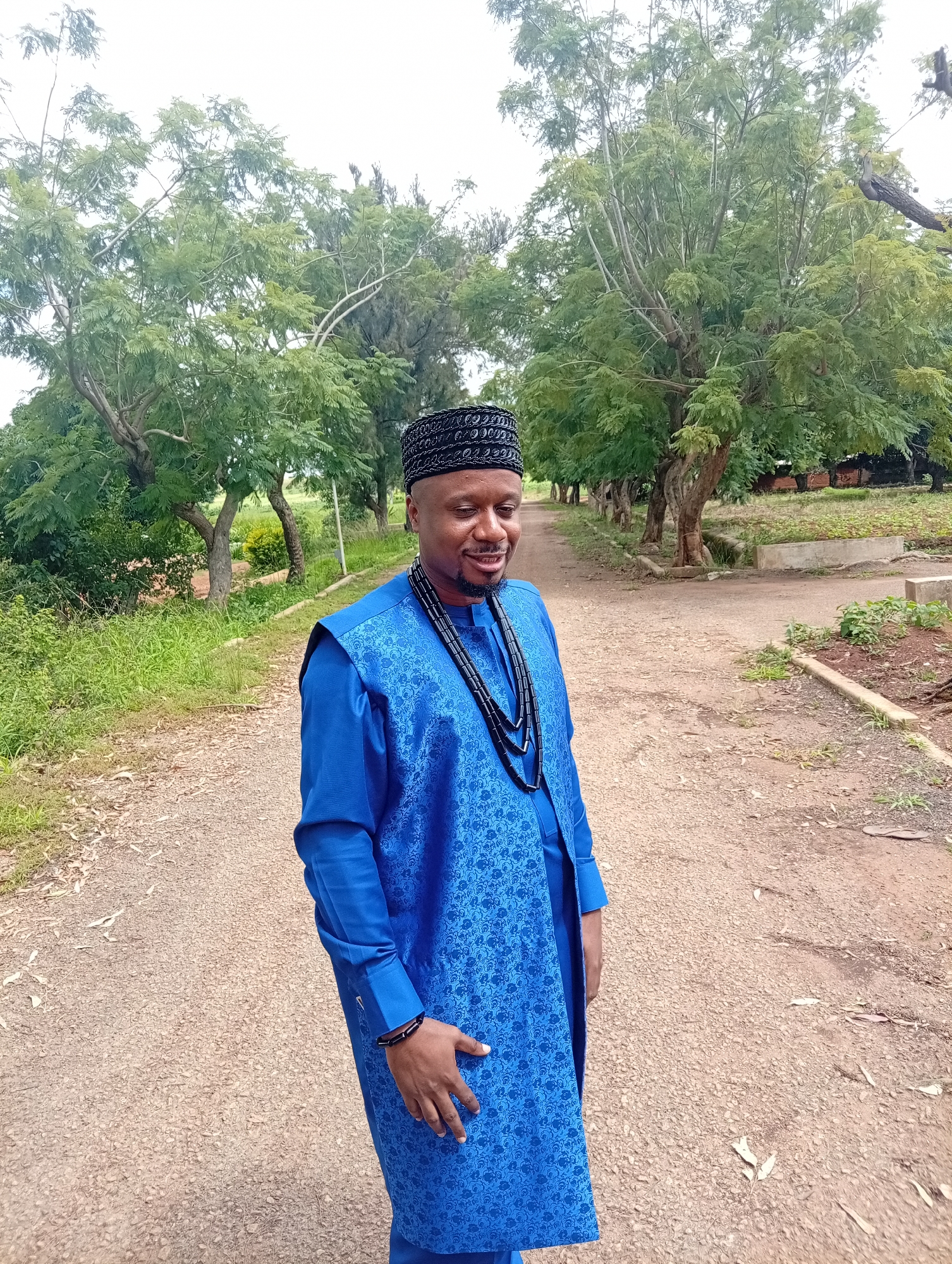 A man wearing a blue traditional outfit and a black patterned cap stands on a dirt path in a grassy area with trees. He is smiling and looking down to his right. The outfit consists of a long-sleeved blue shirt and a blue vest with a patterned design. He is also wearing a black beaded necklace and a black beaded bracelet on his left wrist. The path appears to be in a rural or suburban setting, possibly a park or a residential area. The background shows lush green trees and vegetation. The location is specified as Tambes Mile Four, Nigeria.