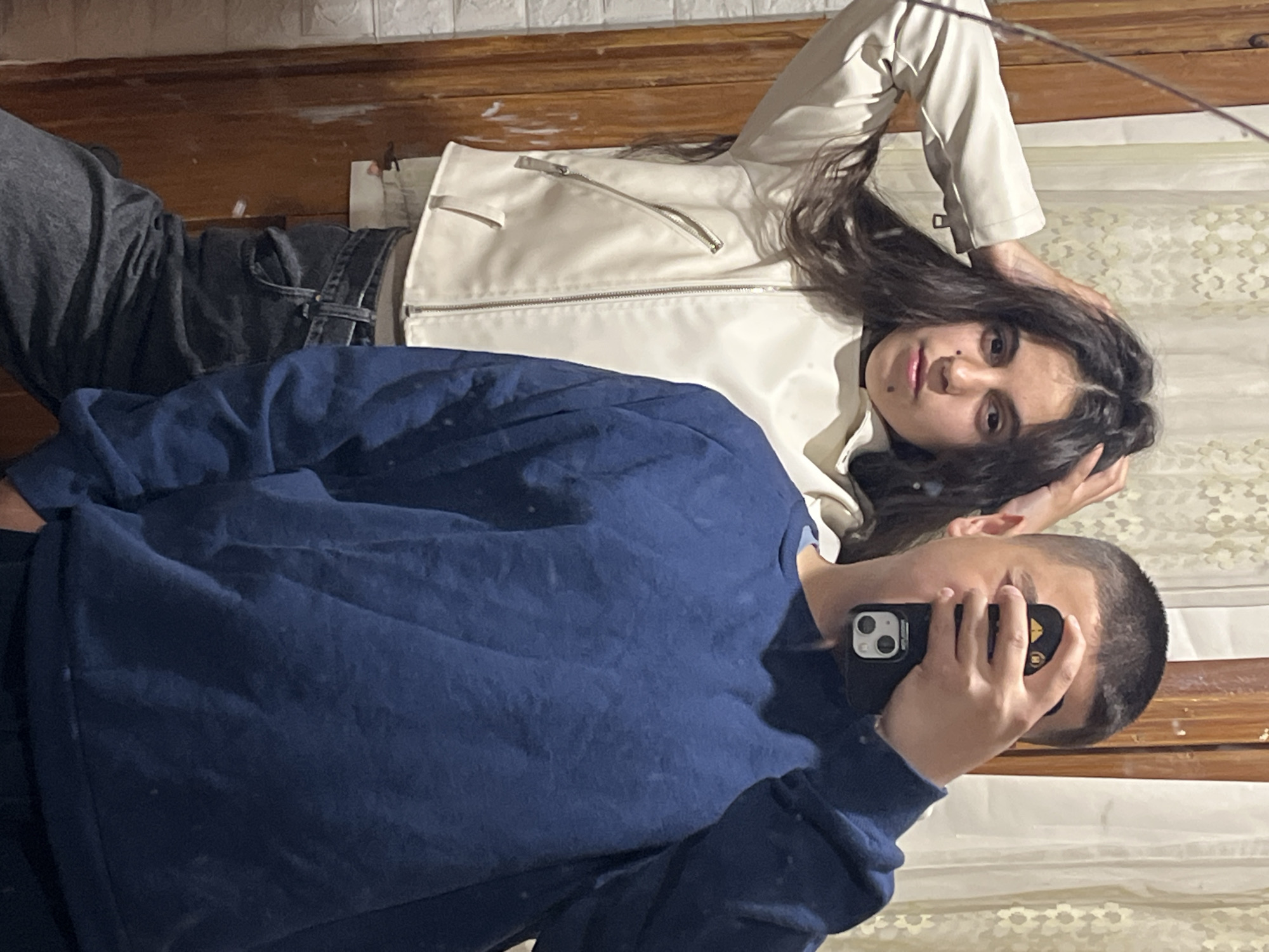 Two people are lying on a wooden floor in an indoor setting. One person is wearing a blue sweater and holding a black smartphone in front of their face. The other person, next to them, is wearing a white jacket and dark jeans. The background includes a textured wall or surface. The image does not provide any specific details to indicate the location is Lozarevo, Bulgaria.
