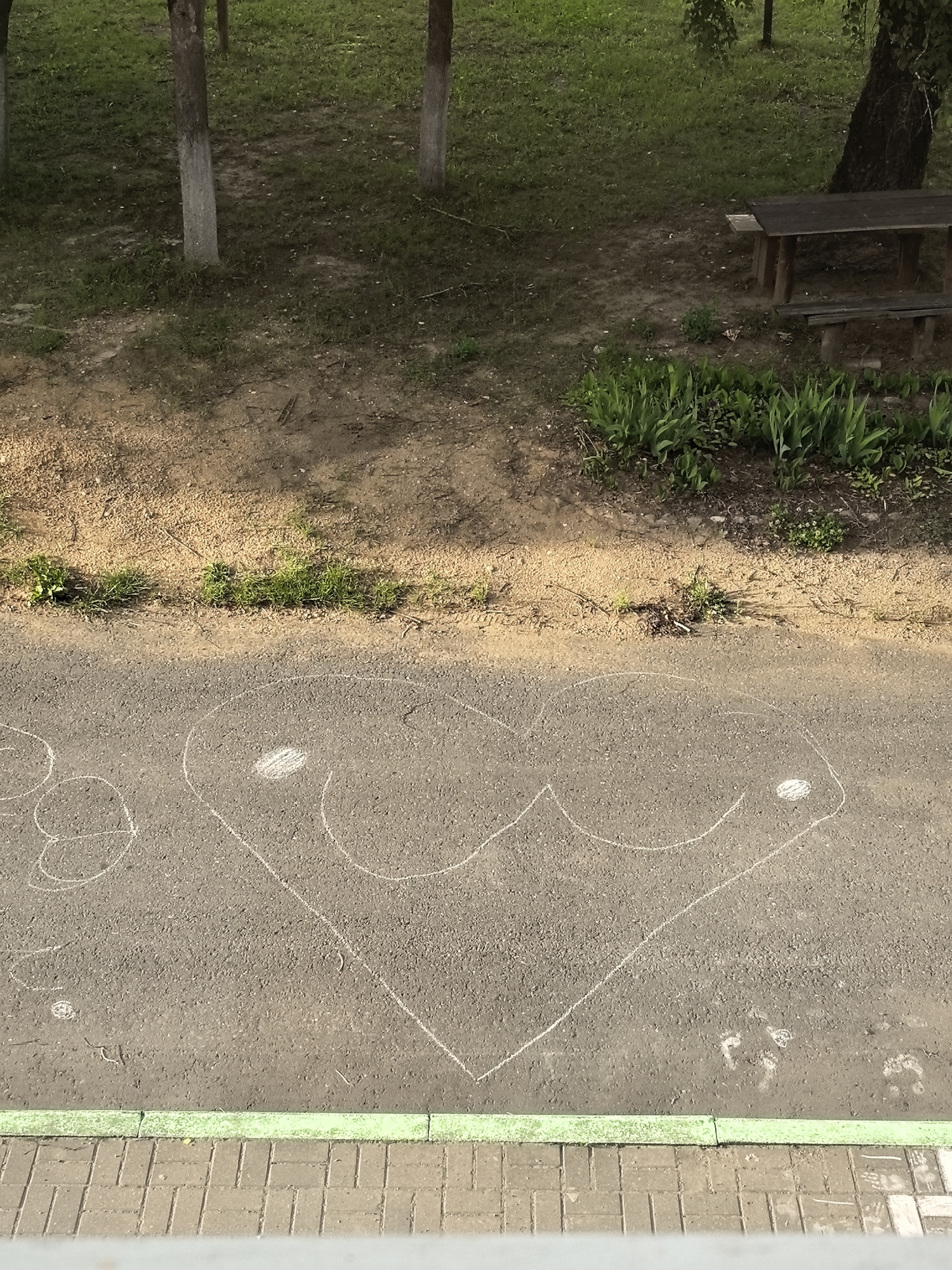An aerial shot shows a chalk drawing of a heart on asphalt. The heart is outlined in white chalk and has two white circles inside it. The heart drawing is on the right side of the asphalt, with a portion of another chalk drawing on the left. The asphalt surface is textured and appears to be a pathway or road. The background consists of a grassy area with trees and a wooden picnic table and benches. A green border and brick pavement are visible at the bottom of the image.