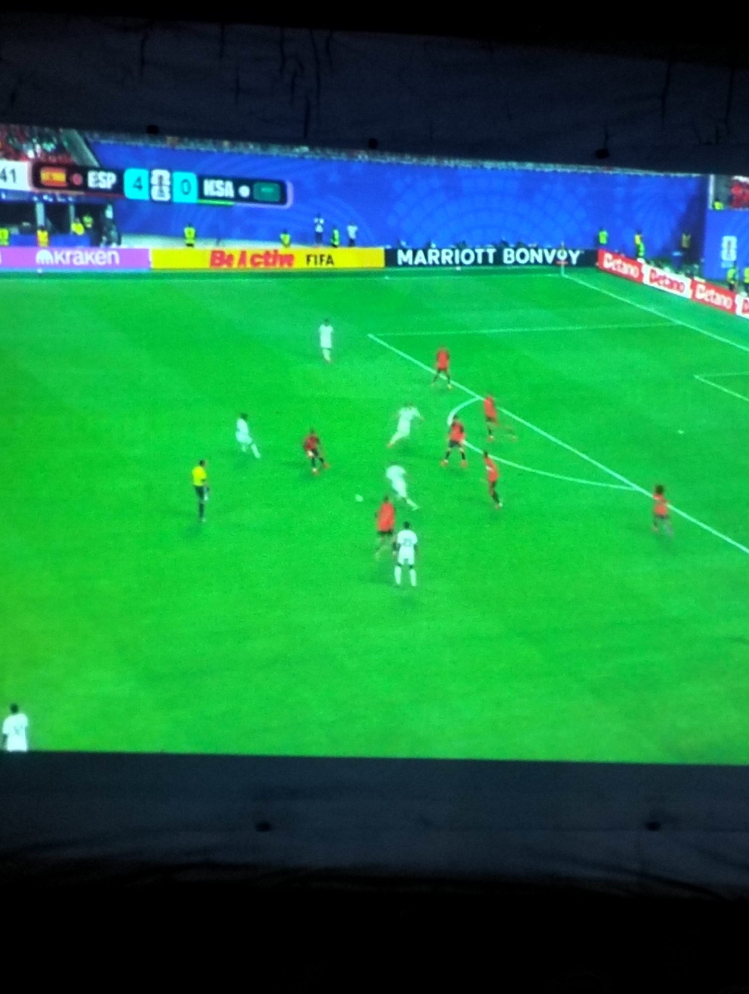 The image depicts a soccer match on a bright green field with a clear blue wall in the background. A scoreboard indicates the score as ESP 4 - 0 KSA. Several players in red and white uniforms are scattered across the field, some in motion, suggesting active play. There are also advertising banners visible along the edge of the field, including "MARRIOTT BONVOY" and "Be Active FIFA". A lone player in a white uniform stands near the bottom left of the frame. The overall scene appears to be a live broadcast of a professional soccer game, possibly in a stadium. There is no indication of the city of Payagaboh, Indonesia in the image.