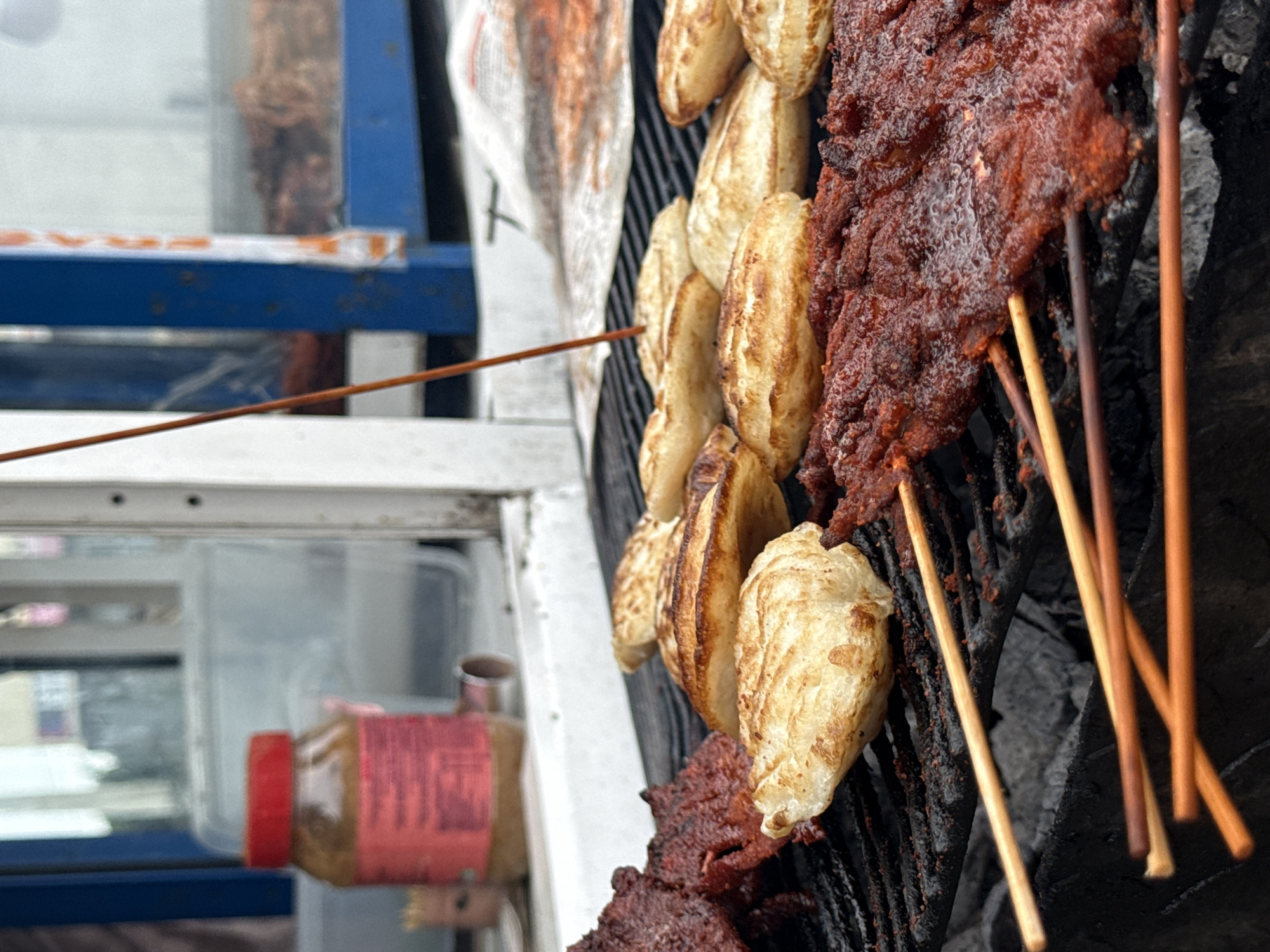 The image shows a close-up, eye-level view of a grill with various food items being cooked. In the foreground, there are several skewers of marinated meat, coated in a reddish-brown spice mixture. The meat appears thin and is being cooked over a charcoal grill. To the right of the meat skewers, there are several round, flattened bread-like items, possibly some kind of flatbread or dough, also grilling on the grate. The grill grate is dark and shows signs of use.
In the background, there is a blurred view of a food stall or window. A red-capped jar, likely containing a condiment or seasoning, is visible on a white surface. Further back, a blue structure with some white and orange accents is present, and through a window or opening in this structure, a glimpse of more food, possibly dried meat, can be seen. The overall impression is that of an outdoor food vendor preparing to serve customers. The setting appears to be an urban environment, possibly a street food stall in Ado, Nigeria, given the context. There are no people clearly visible in the frame, and the focus is entirely on the food and the cooking process.