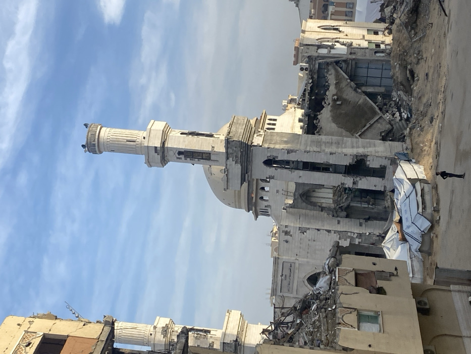 The image depicts extensive urban destruction in Khan Yunis, Palestinian Territory, under a partly cloudy daytime sky. Central to the scene is a severely damaged mosque featuring a light-colored minaret and an intact dome. The minaret shows structural damage and discoloration, with birds perched on its top platform. The main mosque structure below the dome is heavily compromised, exhibiting collapsed walls, exposed interior sections, and broken arched windows. A second, partially obscured and damaged minaret stands to the far left, connected to a ruined multi-story building.
The foreground and midground are covered in large piles of rubble and debris from demolished structures. Amidst this debris, several white and blue tarpaulin-covered objects, possibly collapsed temporary shelters or aid supplies, are visible. A lone young male, dressed in dark clothing, stands upright in the lower center foreground, facing towards the destroyed mosque with his back partially to the viewer, observing the scene. The overall environment is characterized by widespread structural collapse and accumulated demolition waste.