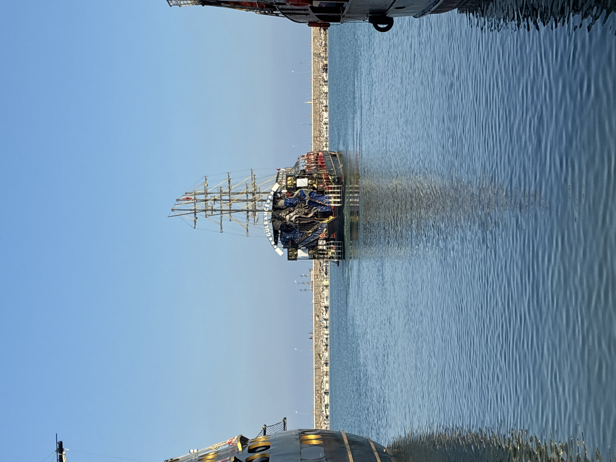 The image shows a view of Alanya Marina in Alanya, Turkey. Three ships are visible. The central ship, a pirate-style ship with multiple masts and flags, is moving away from the viewer towards a breakwater. The breakwater is lined with smaller boats moored in a harbor. Two other ships, one on the left and one on the right, are partially visible and appear to be docked or stationary. The sky is clear and blue, and the water is calm with gentle ripples. The scene depicts a bright, sunny day at a bustling Mediterranean port.