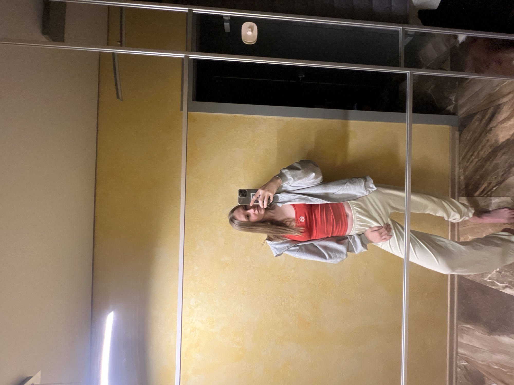 A full-length selfie of a young woman in a gray hoodie, red tank top, and light yellow pants is reflected in a closet mirror. She is holding a smartphone to take the picture. The background includes a textured yellow wall, a black storage space above the closet, and a marble floor pattern to the right of the mirror. The image does not provide sufficient information to determine the location in Khimki, Russia.