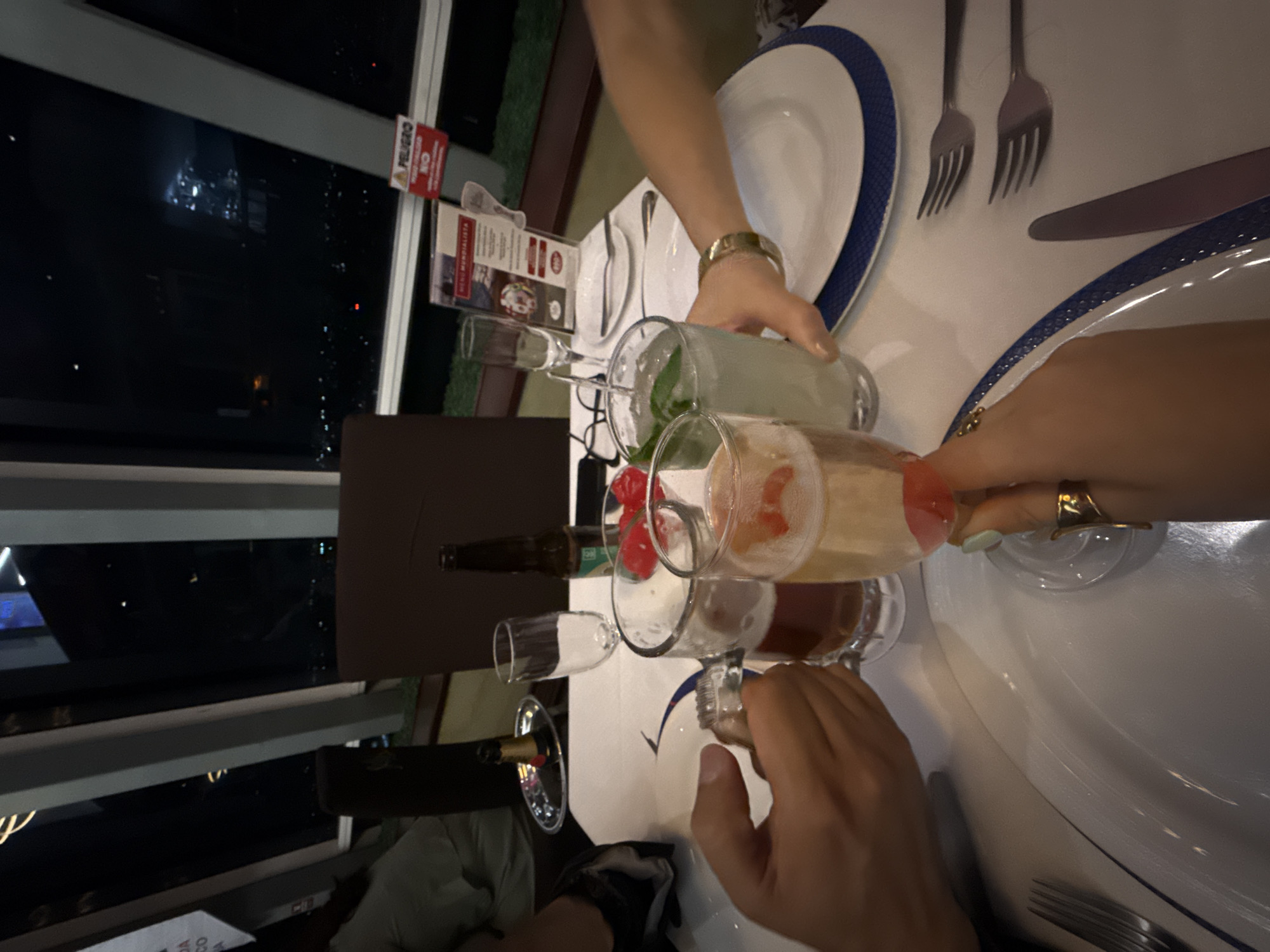 Scene: A dinner table is set for a meal at a restaurant in Mexico City, Mexico, at night. Outside the window, city lights can be seen through the panes.
People: Several individuals are gathered around the table, raising glasses for a toast. Hands are visible holding the glasses, with at least one person wearing a gold watch and another wearing a gold ring.
Subjects: Cocktails and alcoholic beverages are the primary subjects of the scene.
Objects: Various glasses filled with different drinks are on the table. There are plates, cutlery, a menu, and bottles of beer and wine.
Actions: People are raising glasses in a toast. One person is pouring from a small bottle into a glass.
Interactions: The main interaction is the toasting of glasses between people.
Environment: The restaurant setting is sophisticated, with city lights visible in the background. The table is well-set with clean linens, plates with blue edges, and cutlery.