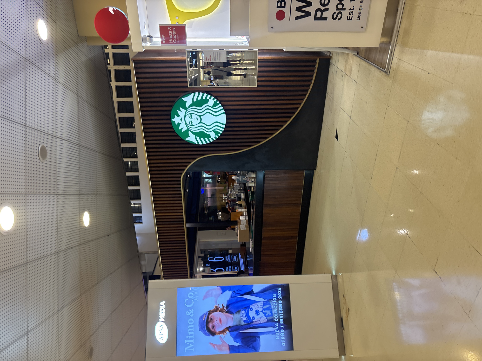 The image shows the front of a Starbucks store inside a building, possibly a mall, in Buenos Aires, Argentina. The store's facade features horizontal wood paneling with a black trim along a curved edge. The Starbucks logo is prominently displayed on the facade. Above the entrance, there is a rectangular sign.
To the left of the Starbucks store, there's a large advertising display showcasing the "Mimo & Co" brand, with text in Spanish indicating a new Autumn/Winter 2026 collection. The display also mentions "APSA MEDIA" and lists pricing details.
The floor is tiled, reflecting light. The area appears well-lit with a combination of natural and artificial lighting. The building has a white ceiling with recessed lights. The overall environment seems modern and clean, indicating a commercial space.