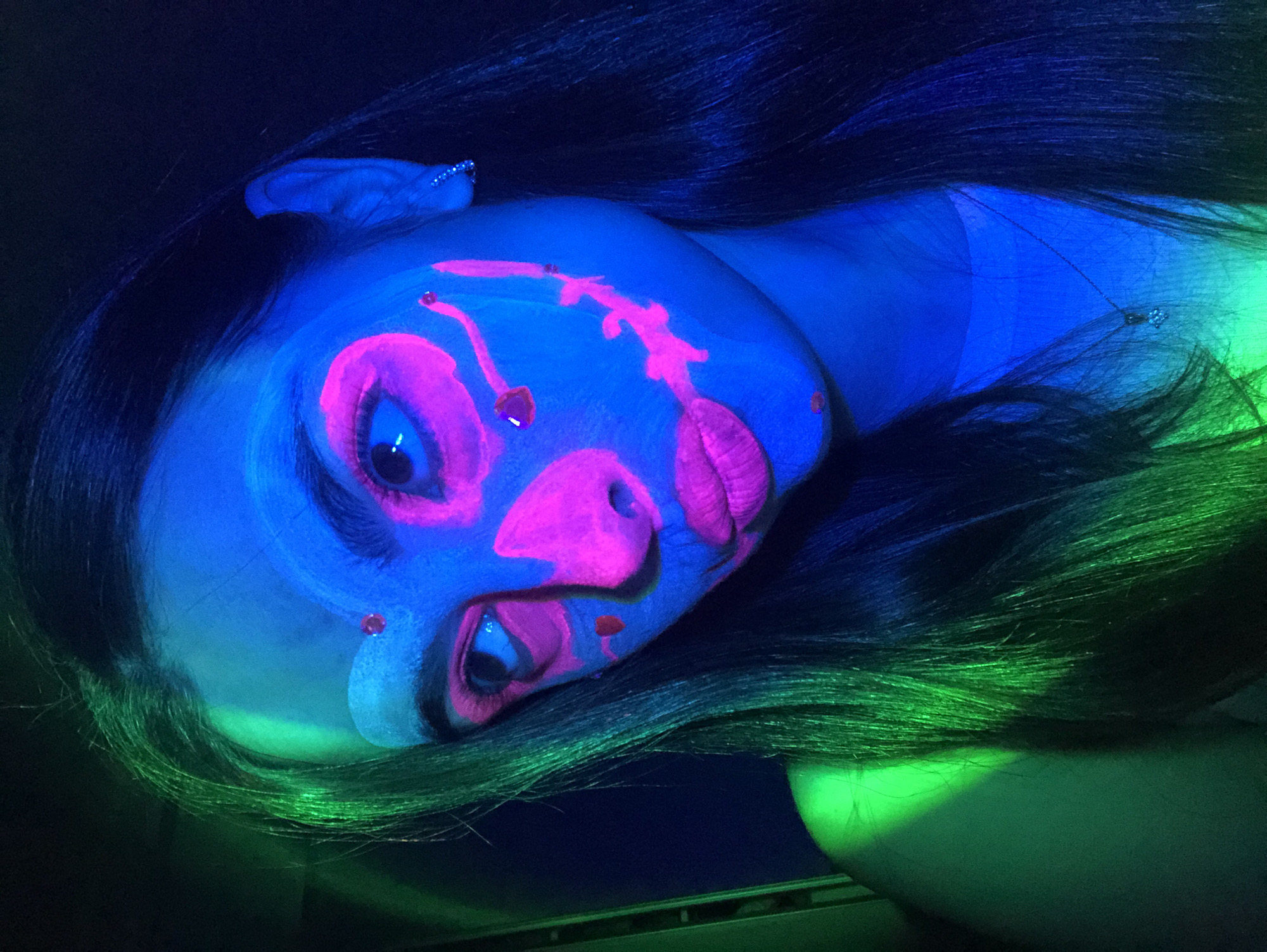 An overhead, close-up shot under UV light features a person with vibrant neon face paint, captured in Noviy, Russia. The individual, likely female, is illuminated by blacklight, causing their skin to glow a striking electric blue and their dark, long hair to show streaks of blue and intense green where the light catches it.
The face is elaborately decorated with fluorescent pink and white paint, creating abstract, artistic patterns. Large pink shapes encircle the eyes, with white accents and dark pupils. The lips are painted a bold neon pink. Intricate pink lines and organic forms extend across the forehead, nose, and cheek, accompanied by small, dark red or orange dots that appear to be part of the design or possibly small embellishments. A subtle shiny earring is visible on the upper part of the left ear, also glowing blue.
The setting appears to be dark, likely an indoor environment such as a club, party, or art event, where blacklight effects are intentionally used. The person's gaze is directed towards the viewer, conveying a sense of artistic presentation or performance. No discernible text is visible in the image.