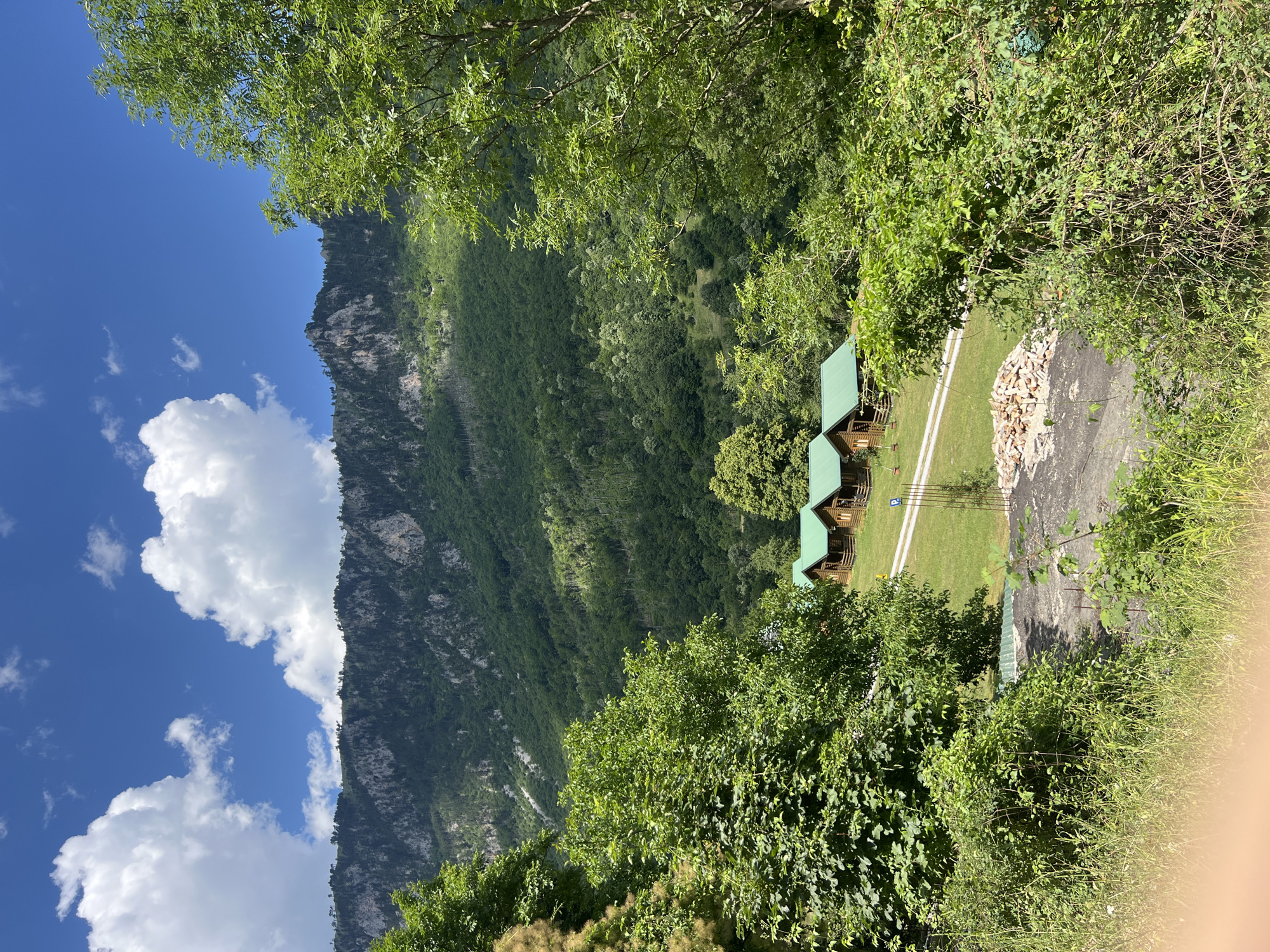 The image depicts a scenic view of a mountainous region with several wooden cabins nestled in a clearing. The cabins, uniformly designed with green roofs, are arranged in a row on a grassy area. A dirt road leads to them. The foreground is dominated by lush green foliage, with trees and bushes obscuring parts of the view. In the background, a majestic mountain range rises, its slopes covered in dense forest. The sky above is a vibrant blue, dotted with fluffy white clouds. The scene suggests a tranquil, rural setting, possibly a resort or a private retreat, in the vicinity of Crna Poda, Montenegro.