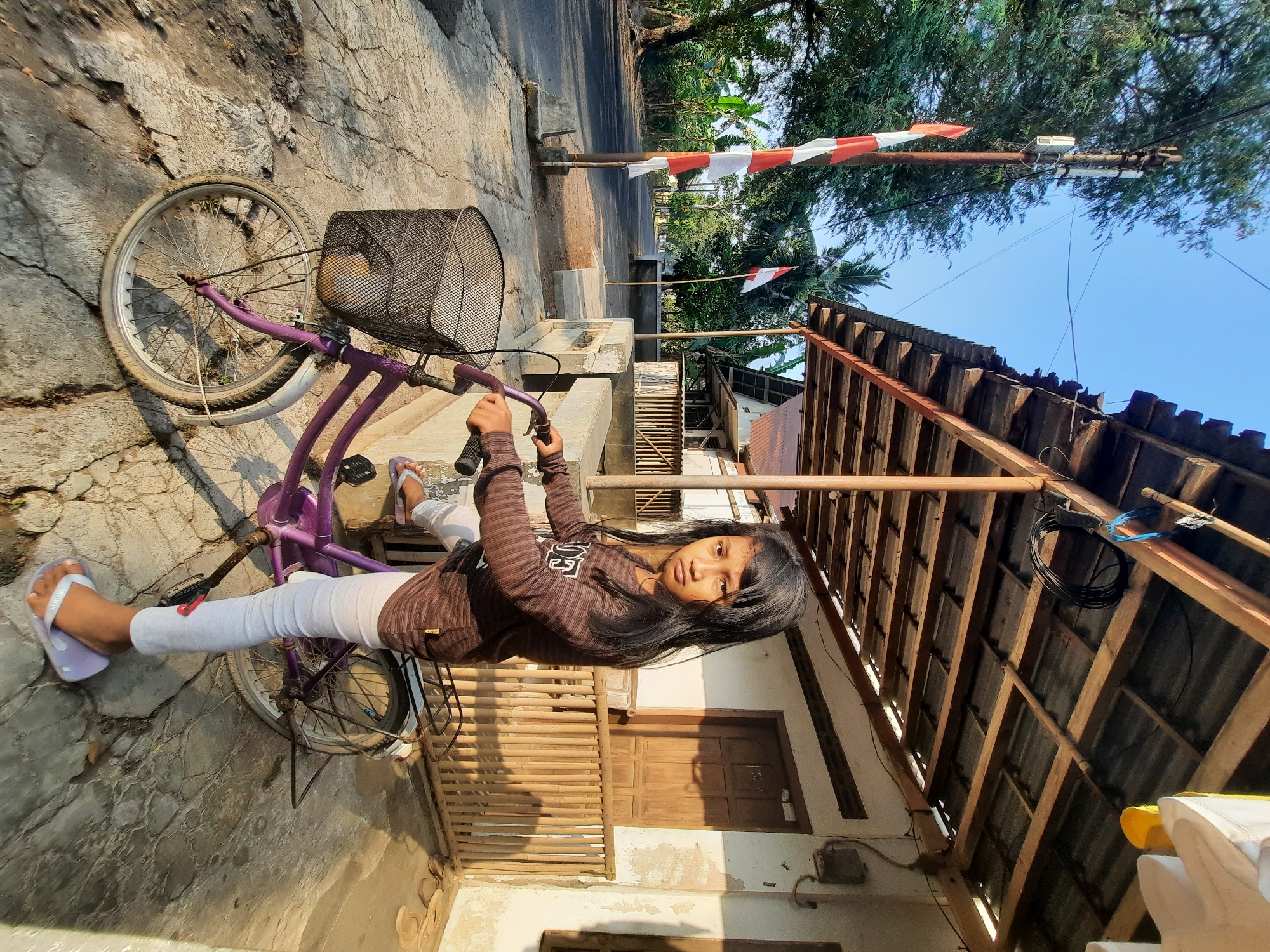 A young Indonesian girl with long, dark hair stands next to a purple bicycle on a cracked concrete sidewalk. She wears a brown striped long-sleeved shirt, white leggings, and light purple flip-flops. The bicycle has a black wire basket in the front. Behind her, a wooden-framed structure with a corrugated roof extends along a building. A wooden fence with vertical bamboo poles is visible on the side of the building. In the background, a paved road runs through a green, leafy environment with trees and houses. Indonesian flags, red and white, are displayed on poles along the road. The scene is illuminated by bright sunlight, casting shadows on the ground. The location is identified as Andong, Indonesia.